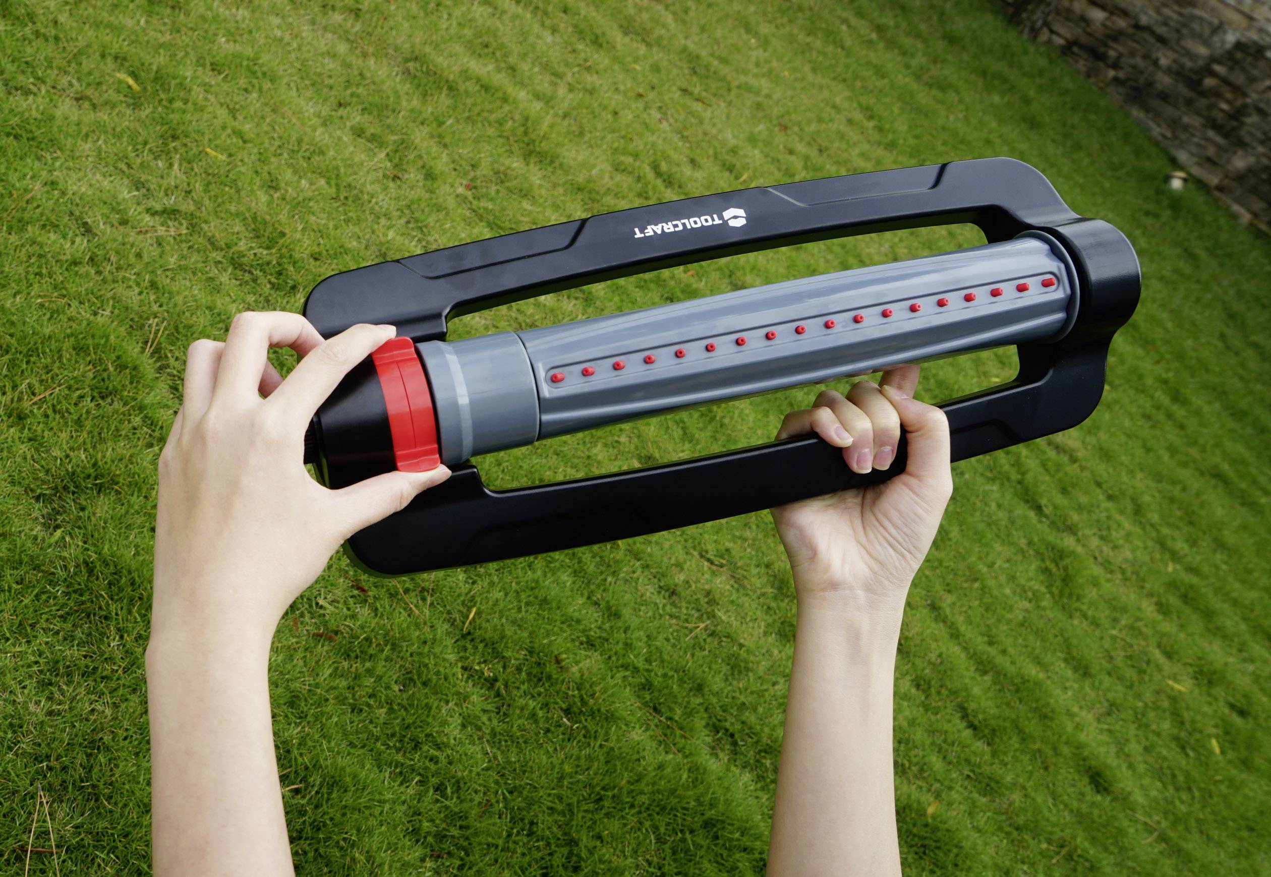 Une personne tient un arroseur rectangulaire à jet réglable dans sa main, alors qu'elle se trouve sur une pelouse verte.