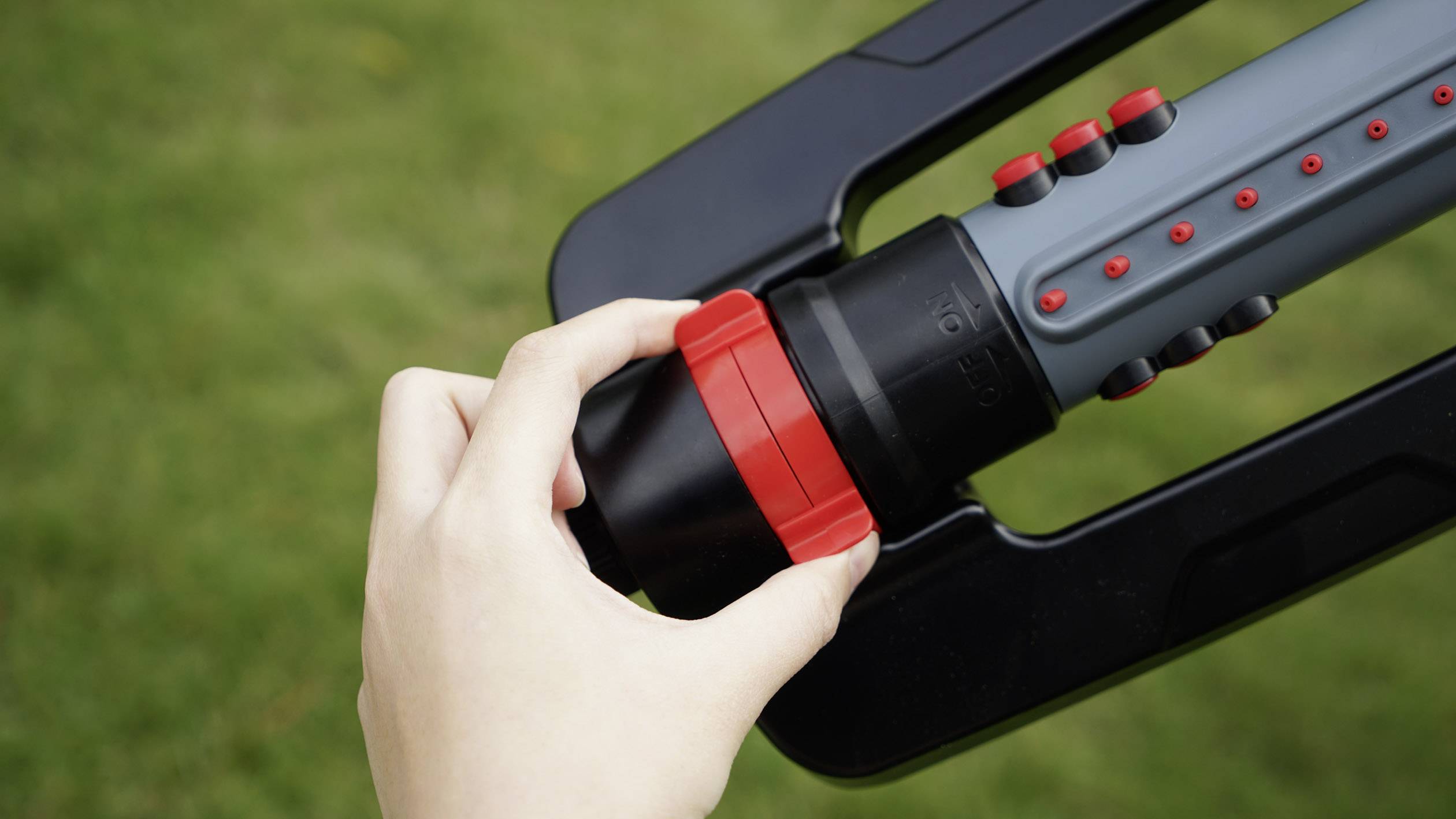 Une main ajuste la valve rouge d'un arroseur de jardin gris sur une pelouse verte.
