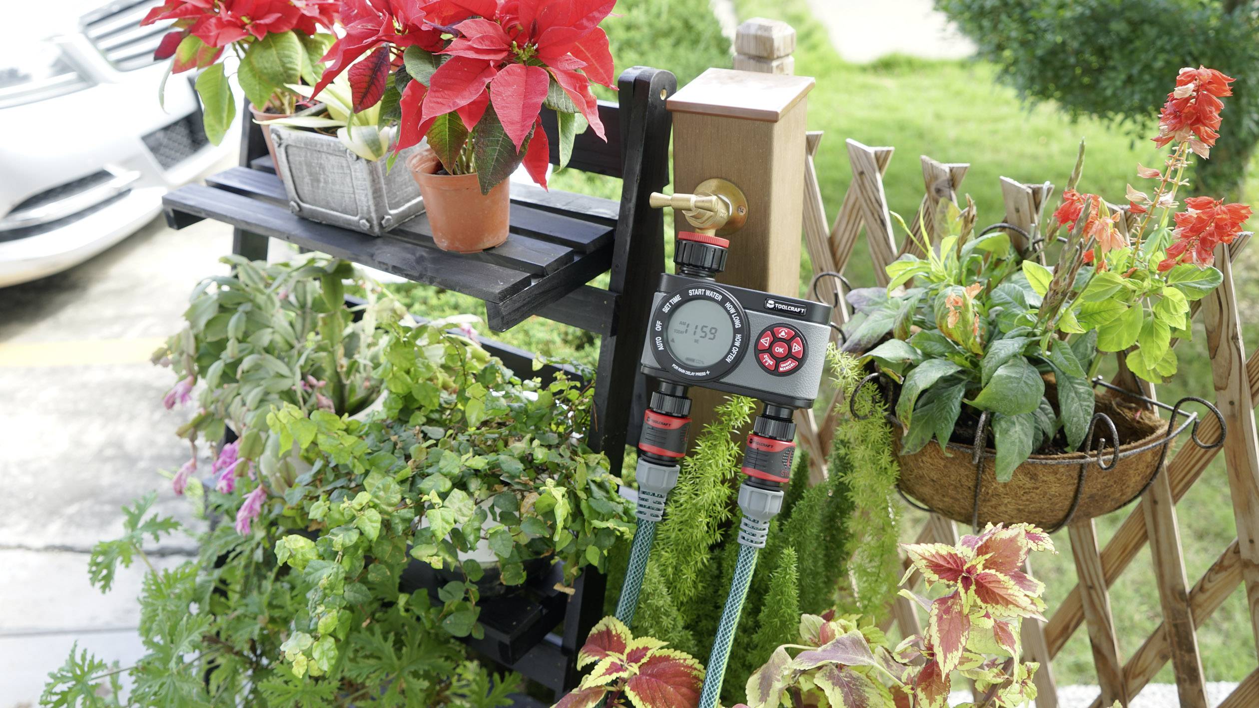 Plusieurs plantes en pots sont disposées sur une étagère à l'extérieur. Un programmateur d'arrosage est fixé à un robinet, entouré de plantes vertes et de fleurs rouges.