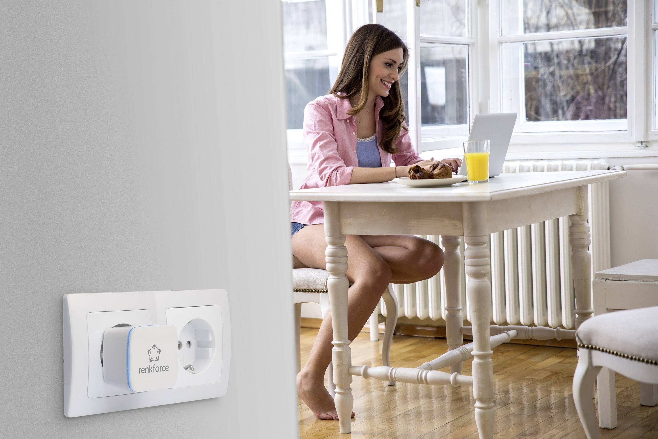 Une femme est assise à une table avec un ordinateur portable, souriante. Au premier plan, une prise électrique avec un adaptateur.
