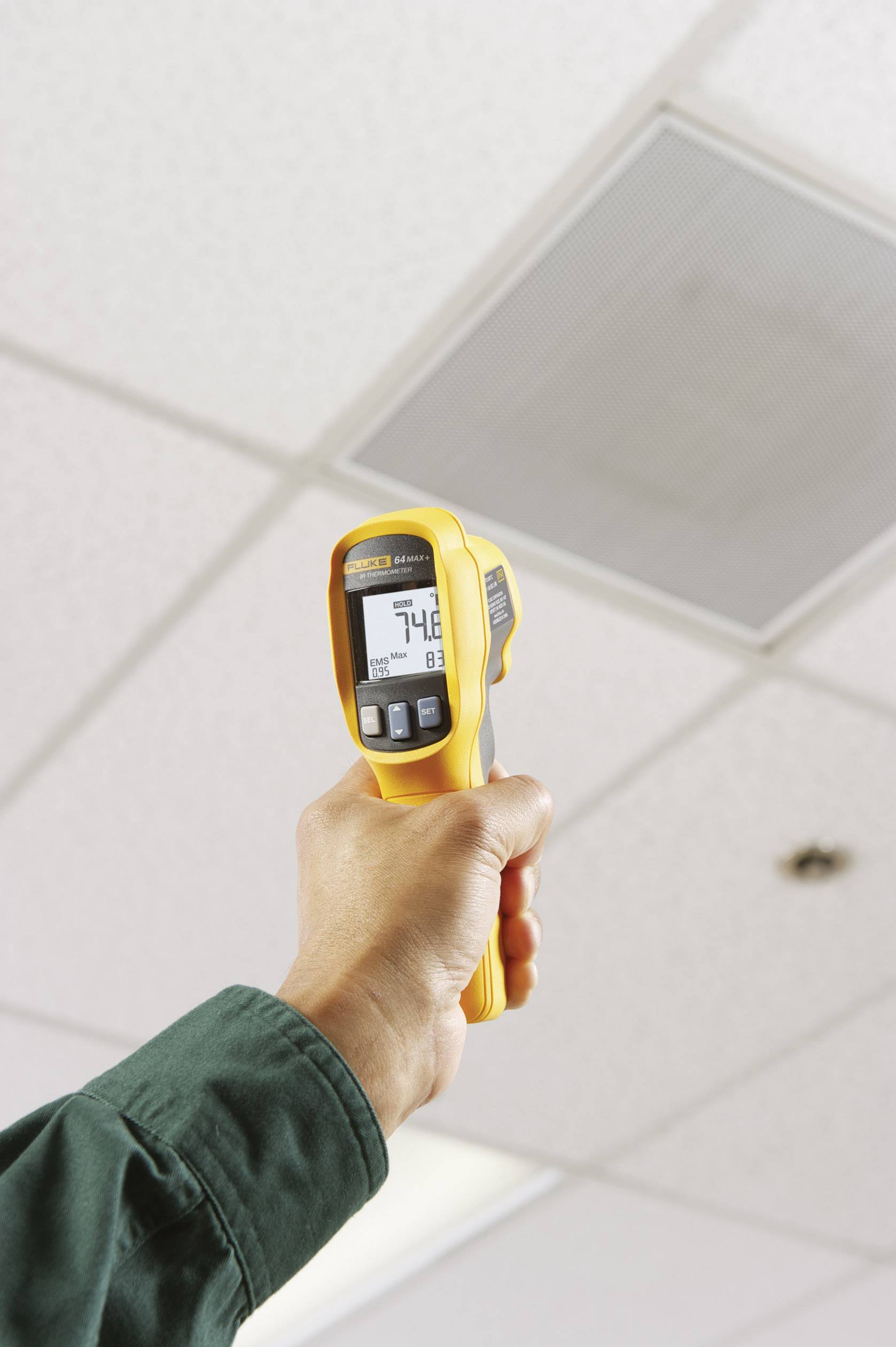 Une personne tient un thermomètre infrarouge jaune et mesure la température de surface du plafond dans un bureau. L'affichage indique « 74,8 °F ».