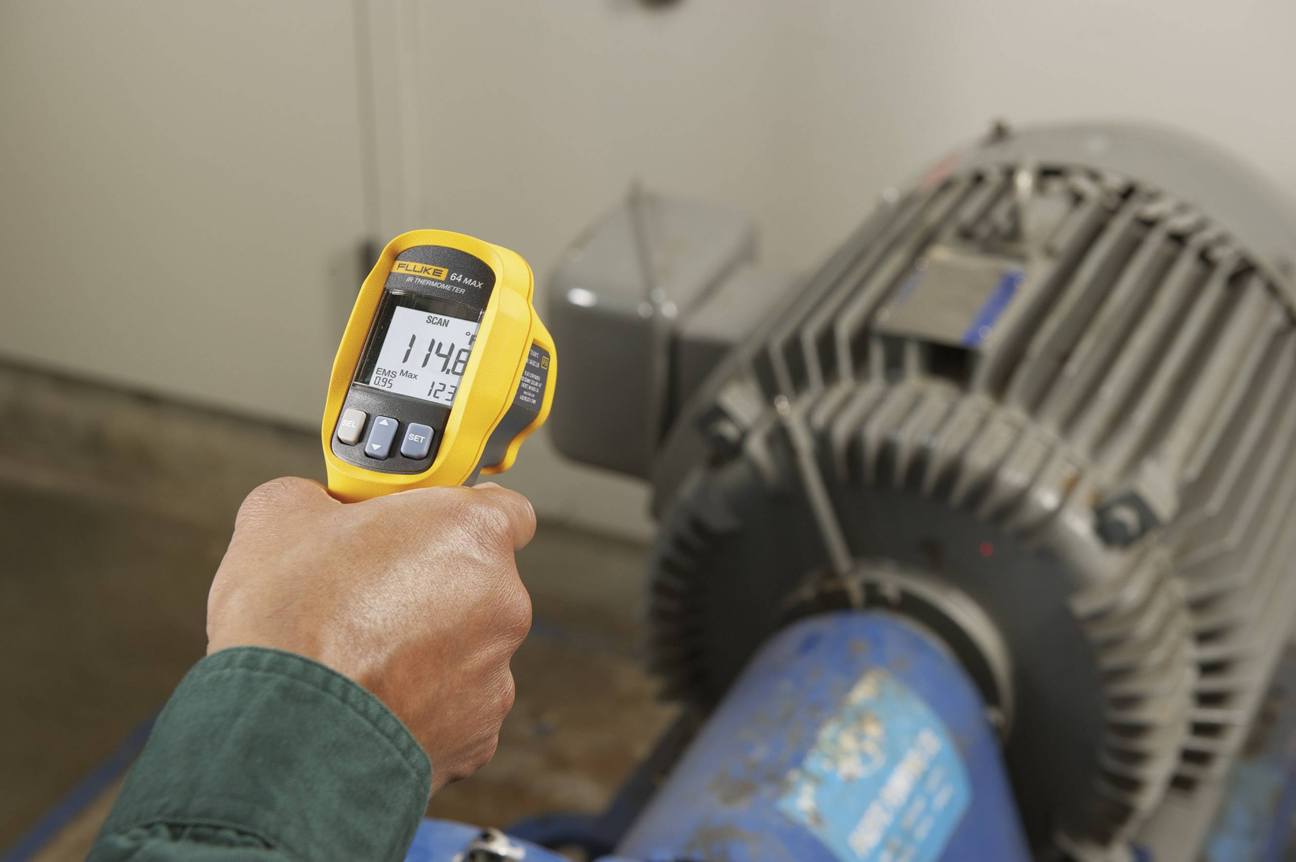 Une personne mesure la température d'une machine à l'aide d'un thermomètre infrarouge jaune. L'affichage indique 114,8 degrés.