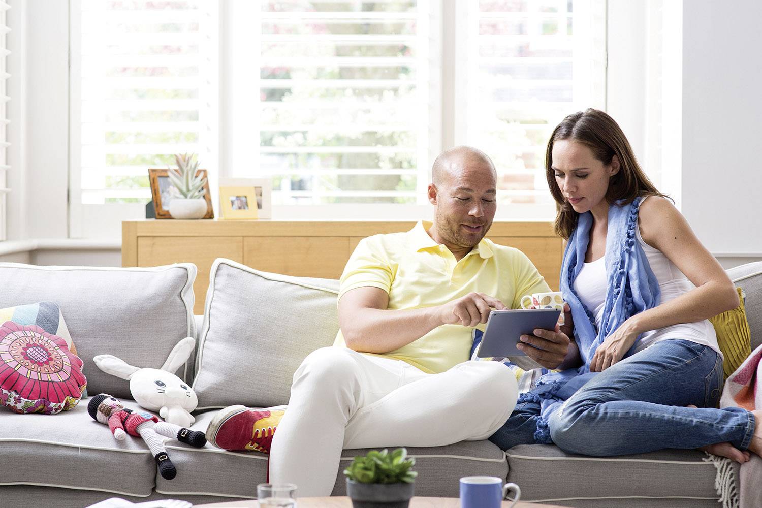 Un couple est assis sur un canapé, regardant ensemble une tablette. Une fenêtre et des décorations sont visibles en arrière-plan.