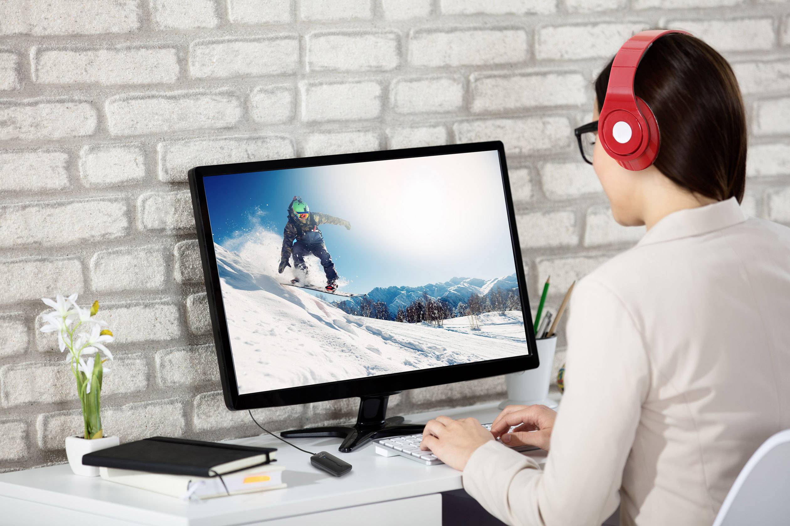 Une personne portant des écouteurs rouges travaille à son bureau et regarde une image à l'écran représentant un snowboardeur dans des montagnes enneigées.