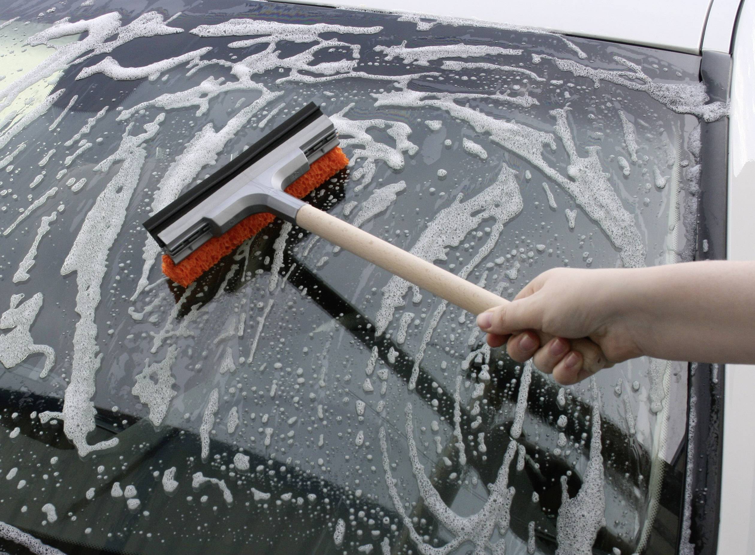 Une main nettoie le pare-brise d'une voiture à l'aide d'un essuie-glace et d'eau savonneuse.