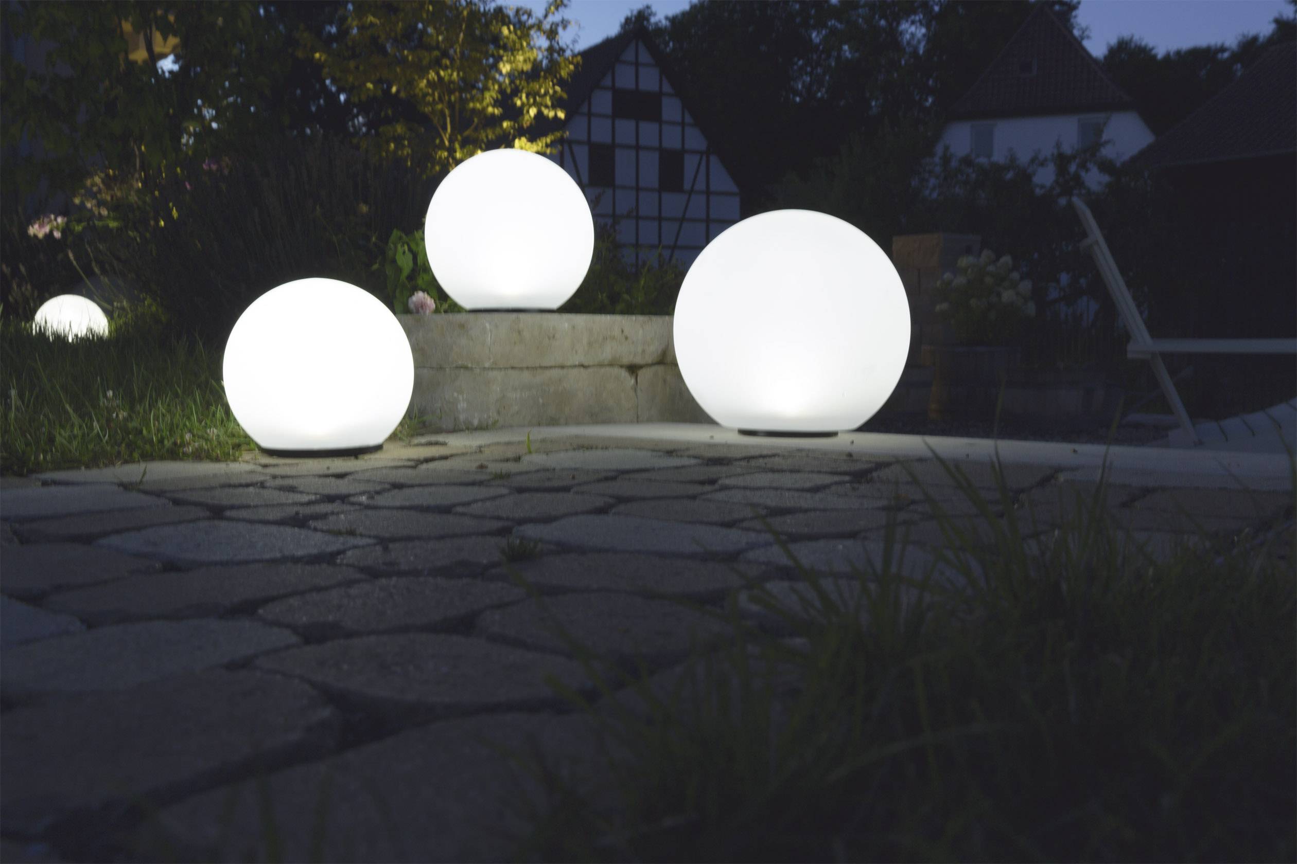 Trois lampes sphériques lumineuses dans le jardin la nuit, entourées de pavés et d'arbres, devant une maison à colombages.