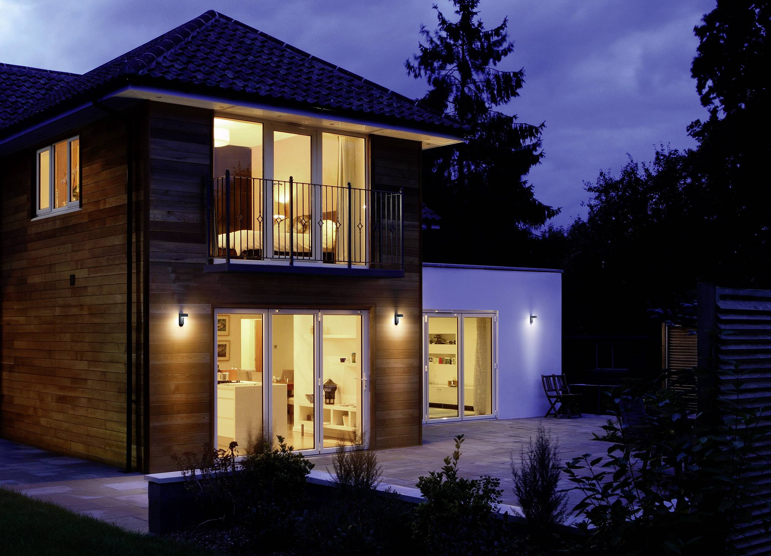 Maison moderne à deux étages avec une grande façade vitrée, illuminée le soir. Environnement avec des arbres et un jardin au crépuscule.