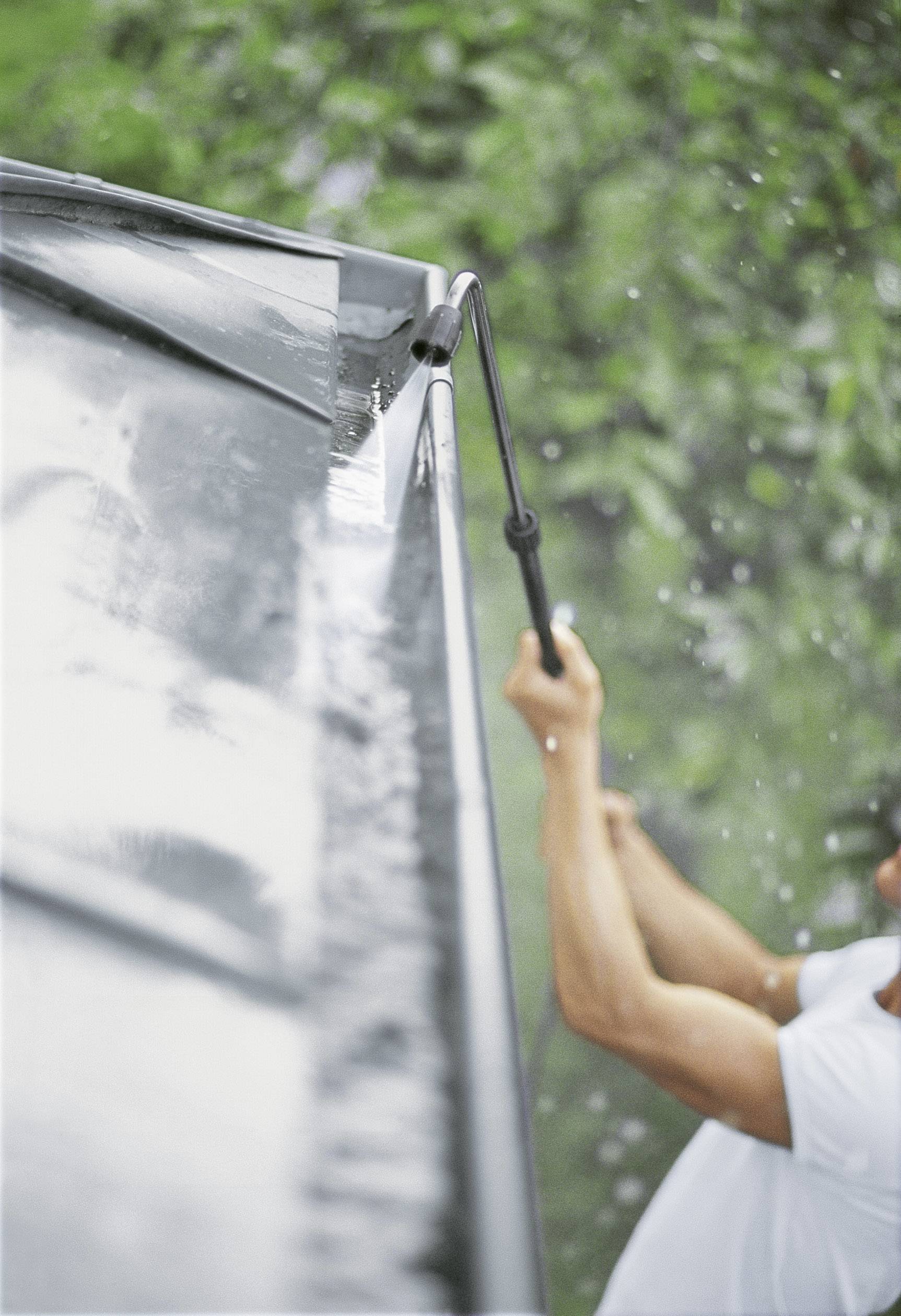 Une personne nettoie le toit d'un bâtiment à l'aide d'un nettoyeur haute pression sous la pluie, avec des arbres visibles en arrière-plan.