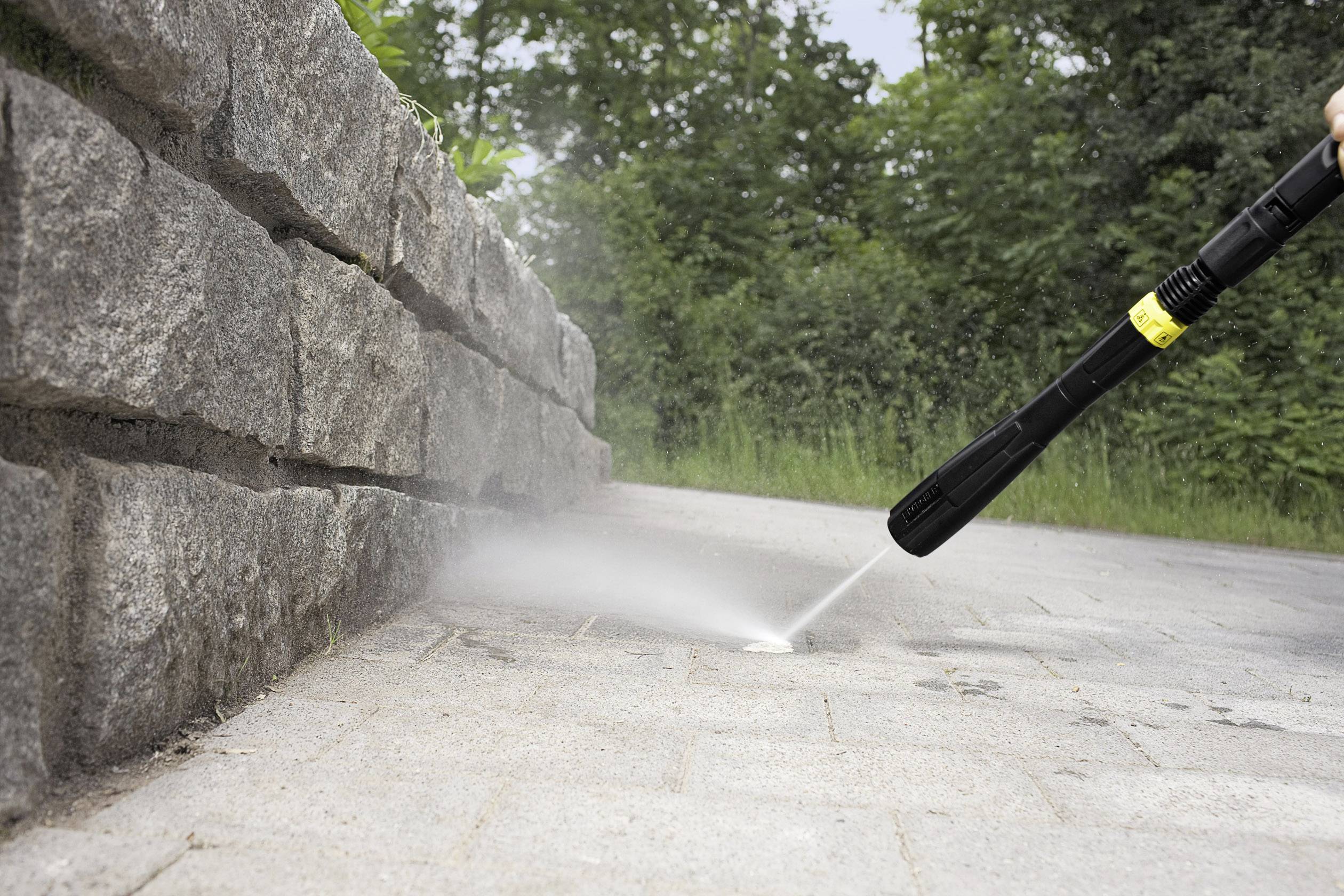 Une personne nettoie les joints d'un chemin pavé à côté d'un mur de pierre à l'aide d'un nettoyeur haute pression à l'extérieur.