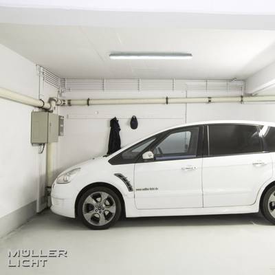 Une voiture blanche est garée dans un garage fermé. Un dispositif d'éclairage se trouve en haut à gauche.