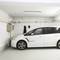 Une voiture blanche est garée dans un garage vide. Deux crochets sont accrochés au mur. Au plafond, un tube fluorescent est installé.