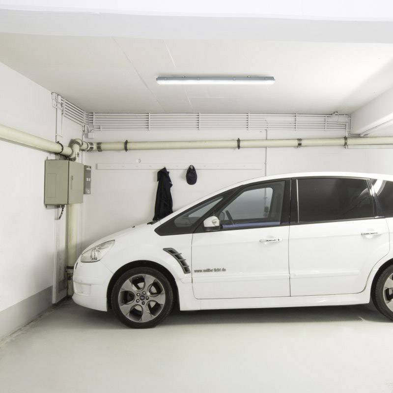 Une voiture blanche est garée dans un garage lumineux. Des tuyaux et un boîtier électrique sont montés sur les murs.