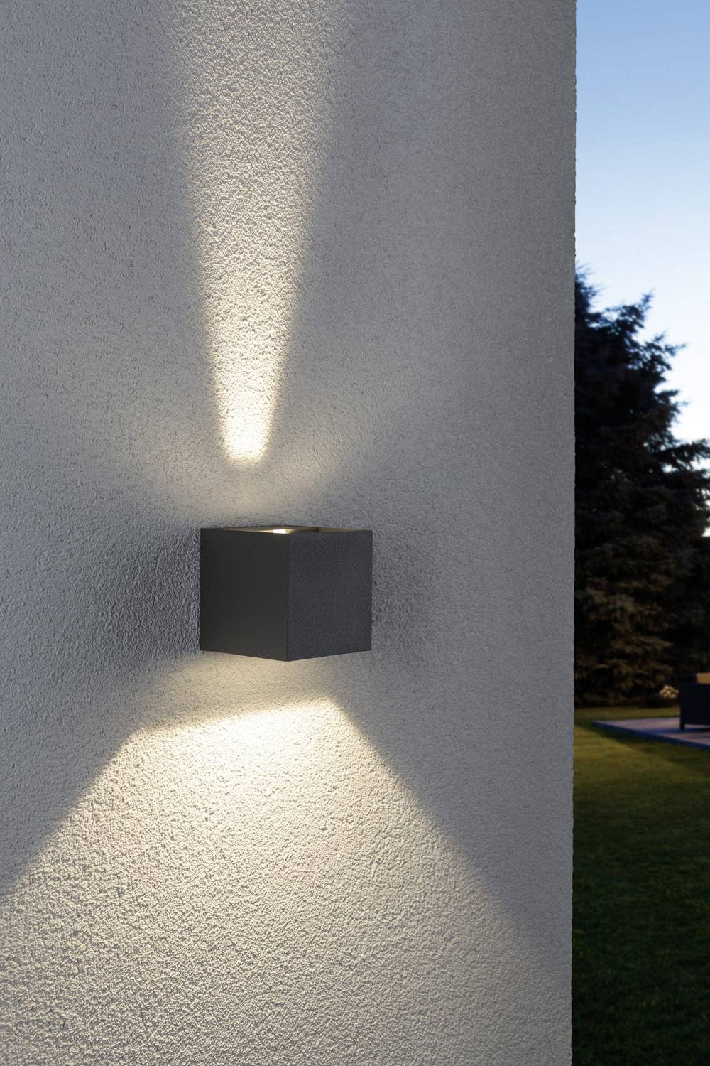 Lampe murale sur un mur extérieur blanc projetant un faisceau lumineux vers le haut et vers le bas. En arrière-plan, des arbres et un ciel bleu au crépuscule.