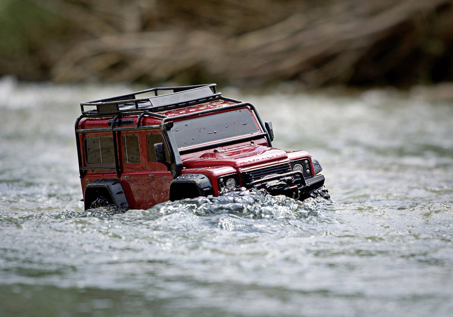 Une petite voiture miniature tout-terrain rouge roule à travers une eau peu profonde, entourée par un cours d'eau en mouvement.