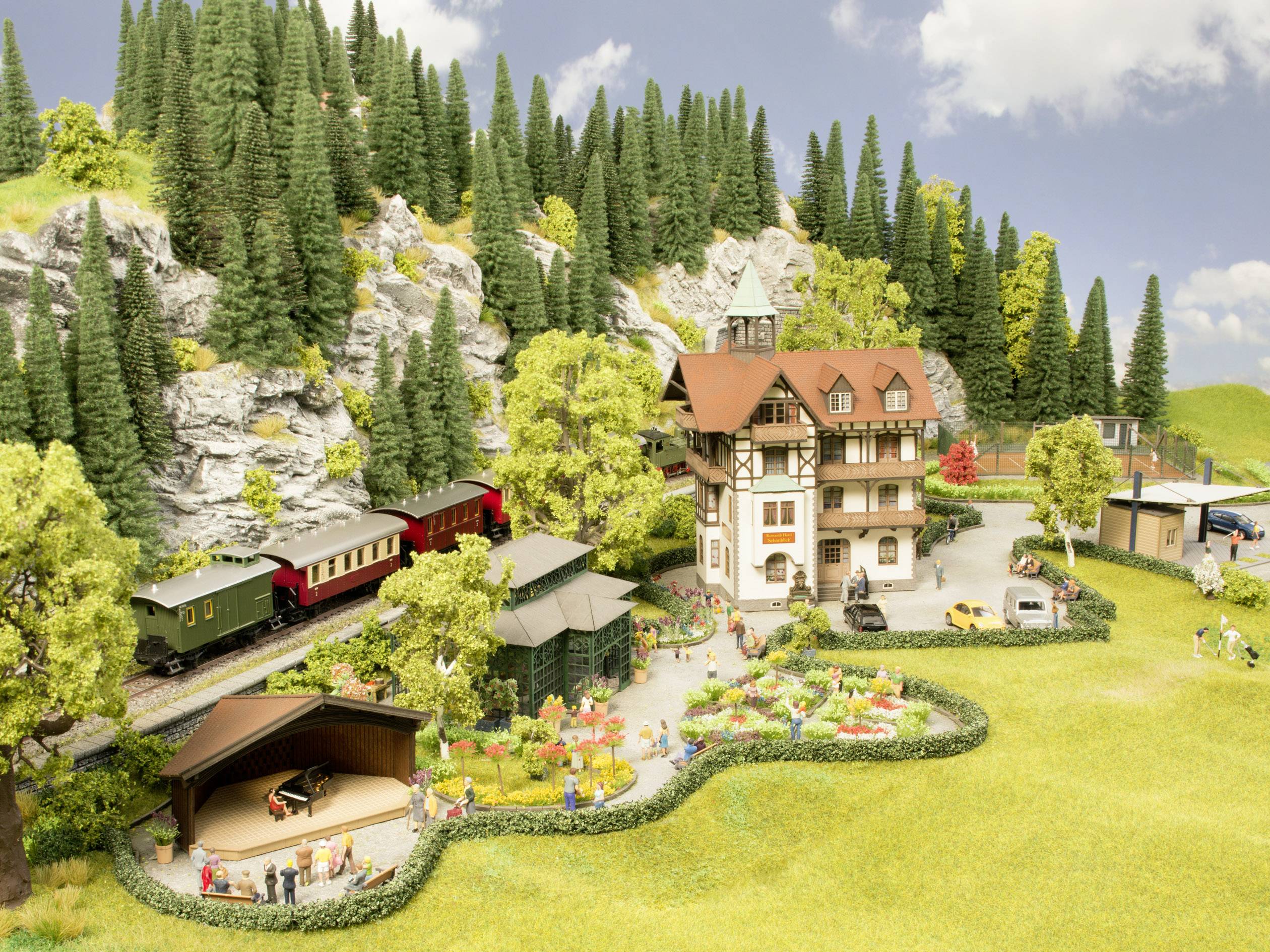 Un modèle d'un paysage avec une maison à colombages, un train en mouvement, des arbres et des personnes lors d'un concert en plein air.