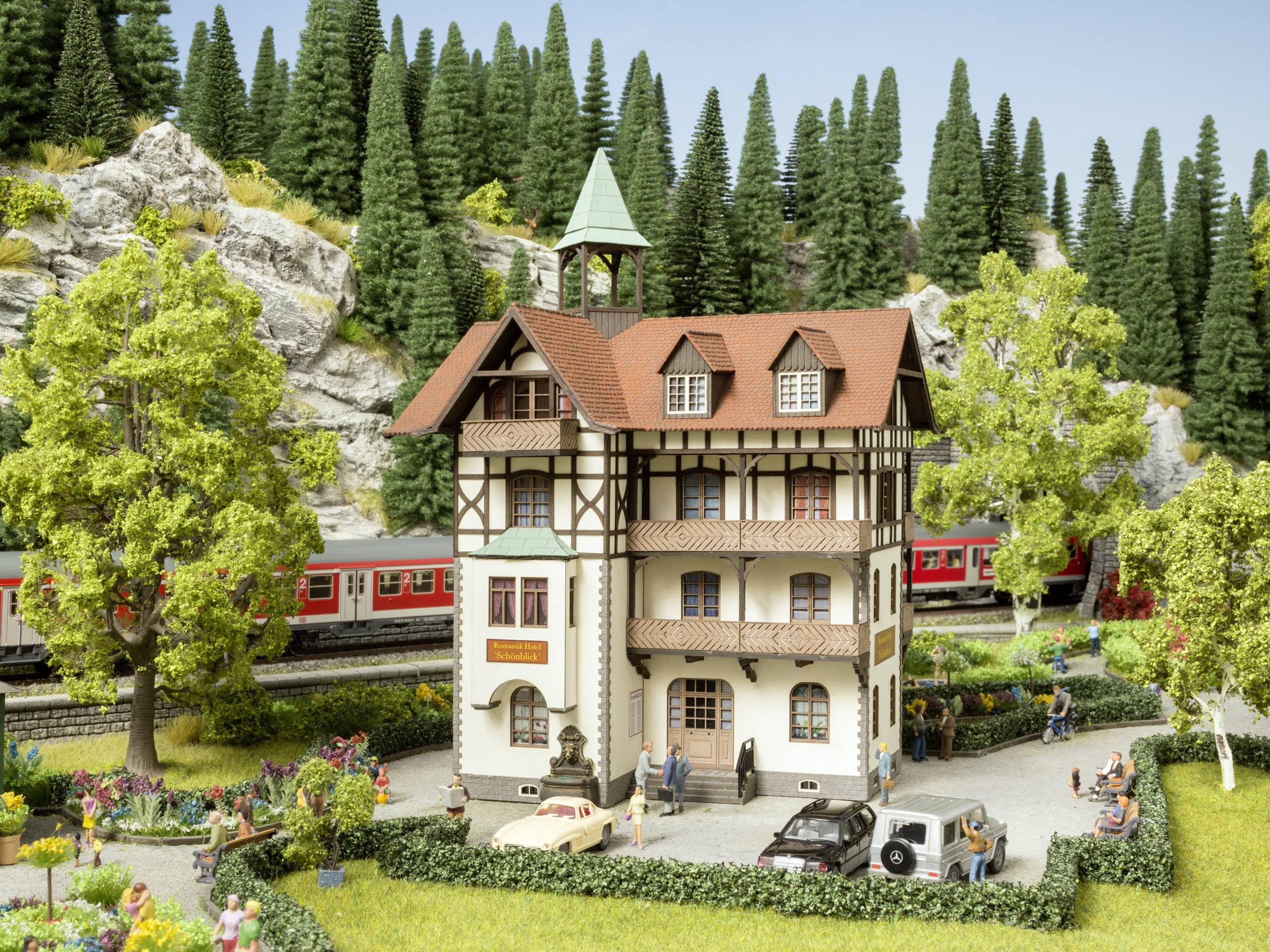 Un modèle réduit représente une maison à colombages sur trois étages avec un jardin, entourée d'arbres et avec un train passant en arrière-plan.