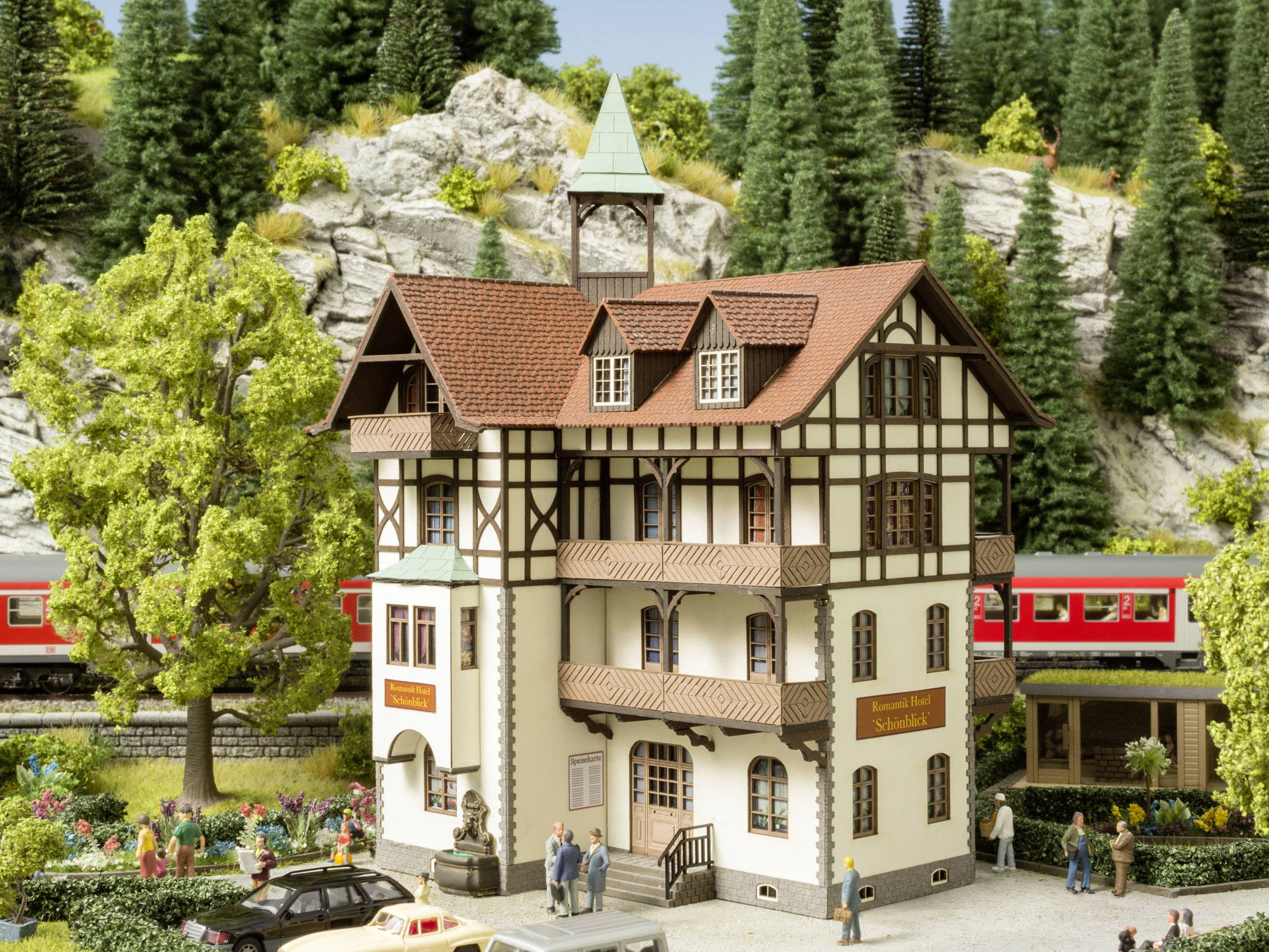 Modèle d'un paysage montagneux avec une maison à colombages, entourée d'arbres et de rochers. Un train rouge passe en arrière-plan.