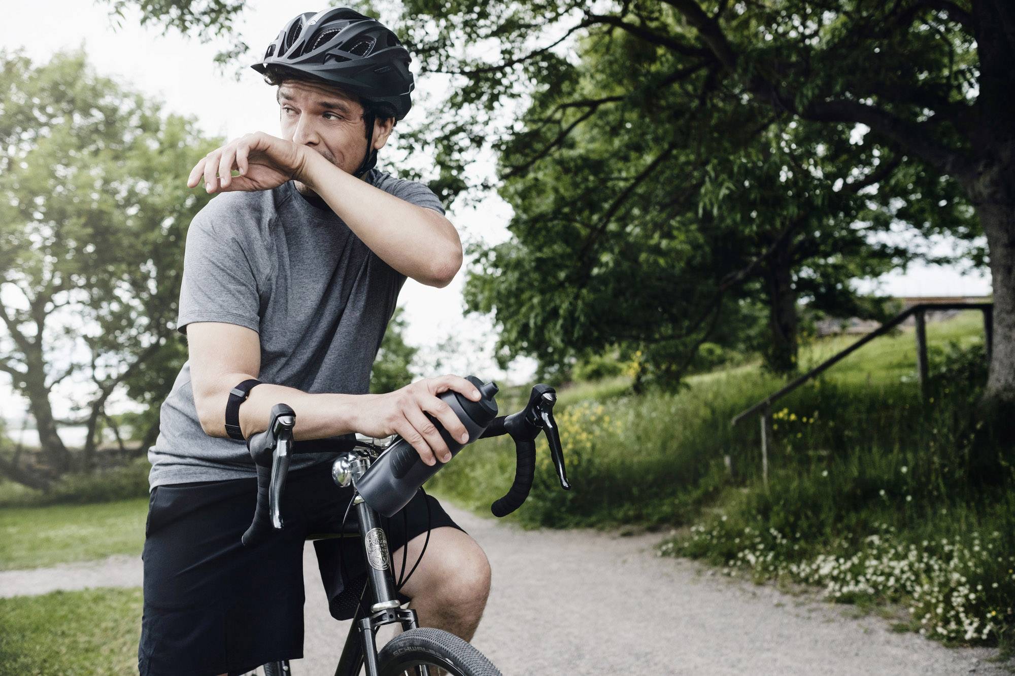 Un homme portant un casque est assis sur un vélo et s'essuie le front de sueur dans un parc ensoleillé.