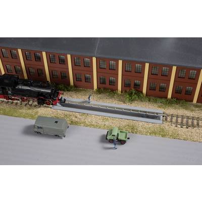 Scène de train miniature : Un train est stationné sur des rails à côté d'un bâtiment en briques. Deux petits véhicules et des figurines sont présents sur la route.