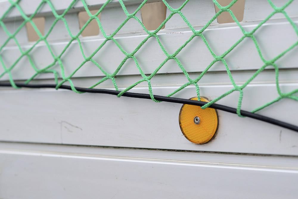 Un filet vert est fixé à une clôture blanche, avec un réflecteur orange et un câble noir en dessous.