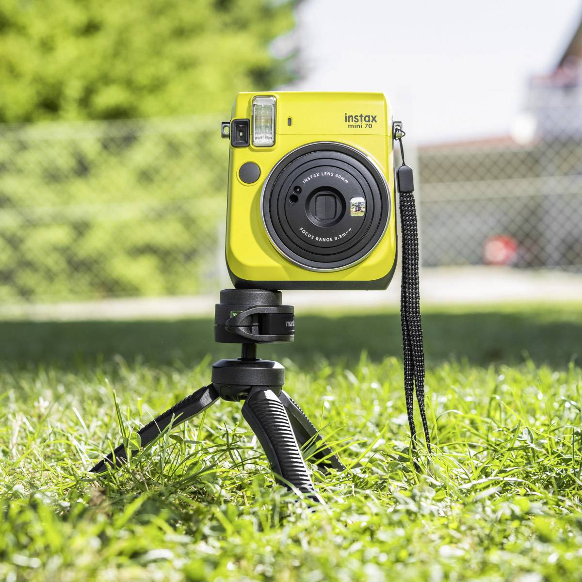 Un appareil photo instantané jaune sur un trépied, positionné sur une prairie verte devant une clôture floue en arrière-plan.