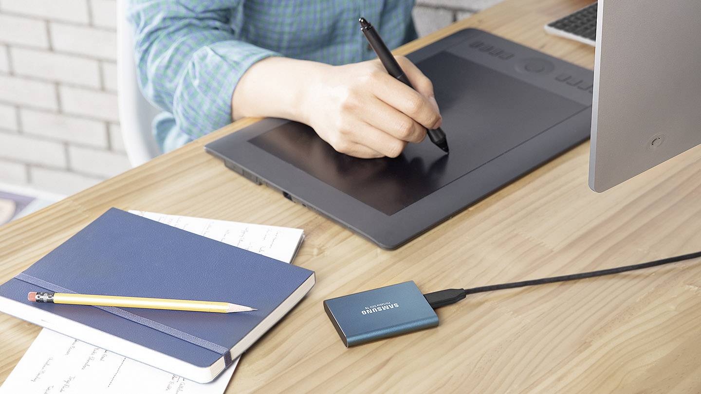 Une personne dessine avec un stylet sur une tablette graphique. À côté d'elle, des cahiers, un crayon et un disque dur externe sont posés sur la table.