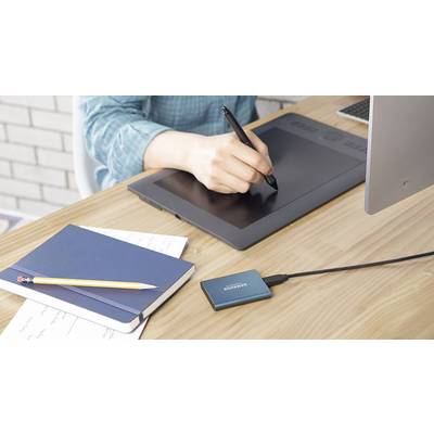 Une personne dessine avec un stylet sur une tablette graphique. À côté d'elle, des cahiers, un crayon et un disque dur externe sont posés sur la table.