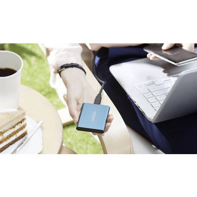 Une personne utilise un ordinateur portable à l'extérieur, tenant un disque dur externe Samsung à la main. Un gâteau et une tasse de café sont posés sur une table.