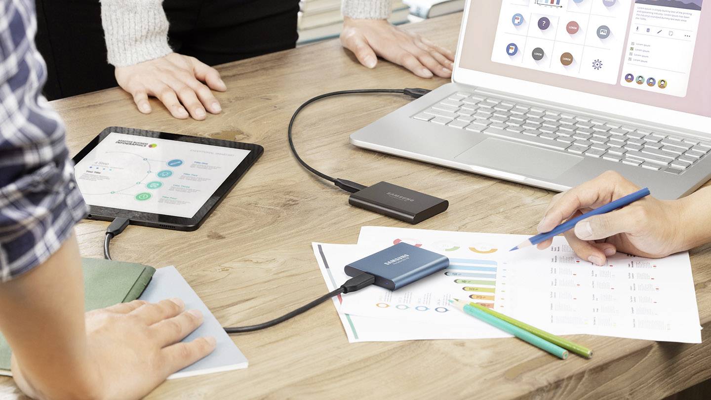 Des personnes sont assises autour d'une table avec un ordinateur portable, une tablette et des diagrammes. Un disque dur externe est connecté. L'accent est mis sur la collaboration et le partage de données.