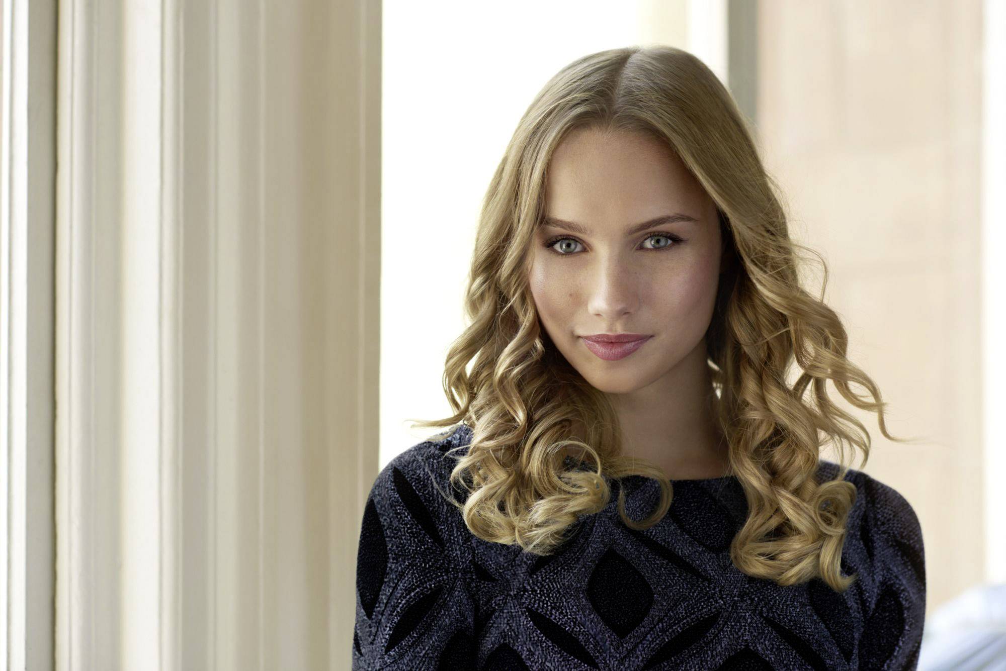 Une personne aux cheveux bouclés blonds se tient devant une fenêtre, portant un pull sombre à motif géométrique et esquissant un léger sourire.