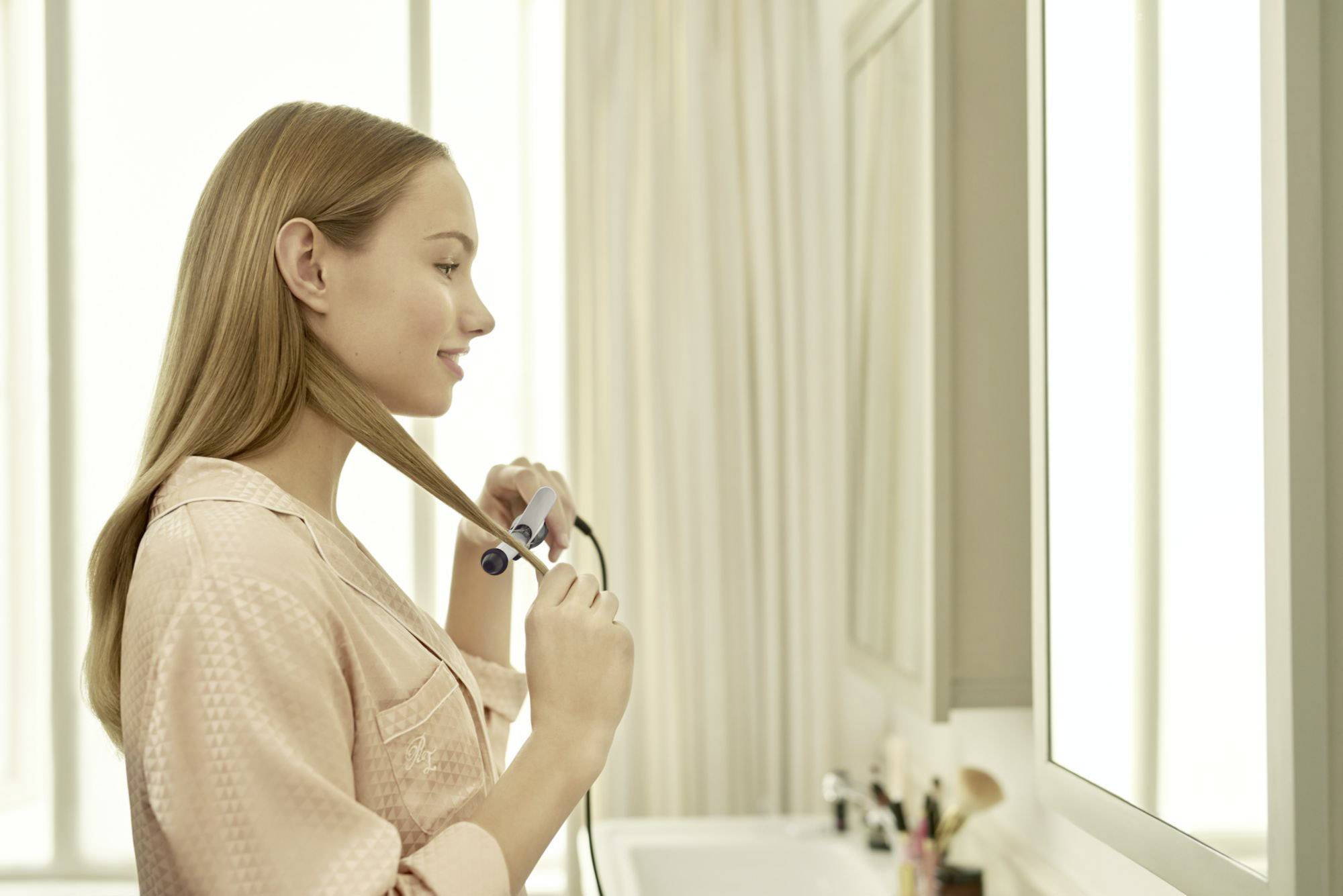 Une femme dans la salle de bain lisse ses cheveux avec un fer à lisser. Elle porte un peignoir rose. Des produits de soin sont visibles en arrière-plan.