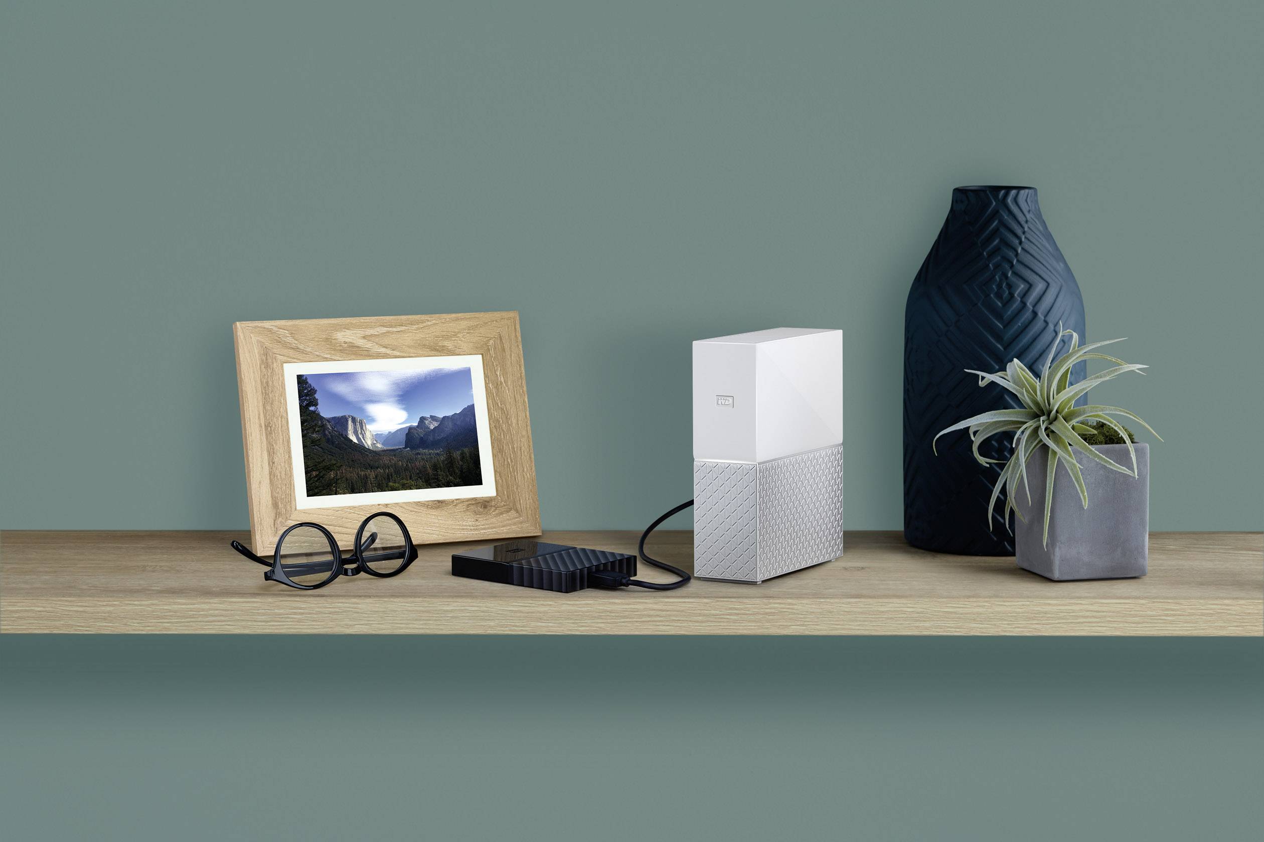 Une étagère en bois moderne contient une photo de paysage encadrée, des lunettes, un disque dur, un routeur blanc et deux vases.