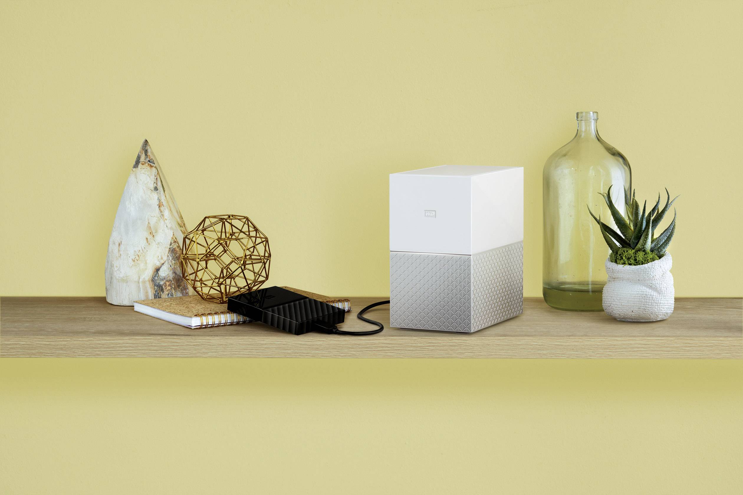 Un bureau décoré, avec un disque dur externe, un smartphone et des plantes devant un mur jaune.