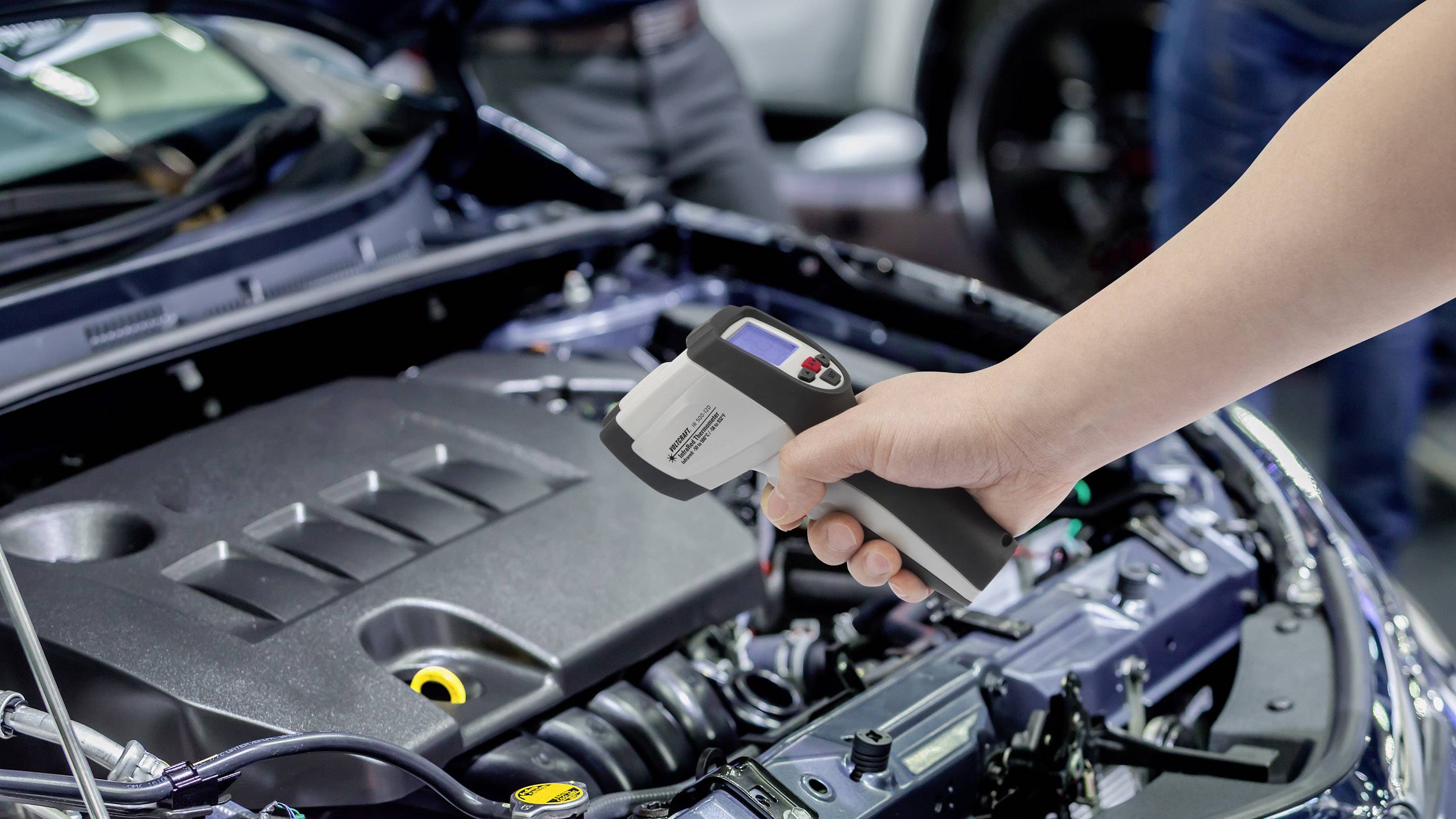 Une main tient un thermomètre infrarouge au-dessus du compartiment moteur ouvert d'une voiture pour en mesurer la température.