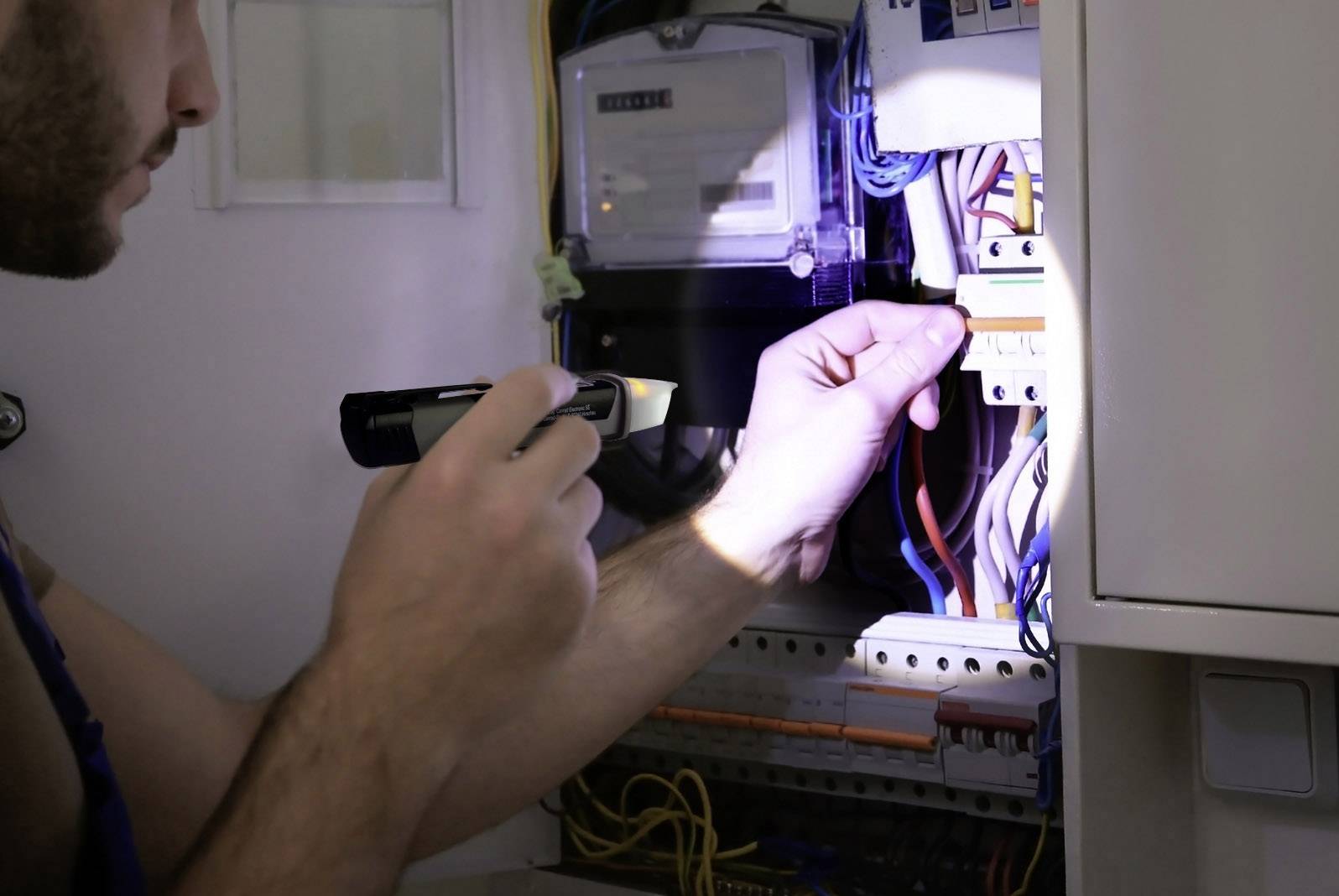 Un électricien vérifie le tableau électrique à l'aide d'une lampe torche, examinant les connexions de câbles et les fusibles.
