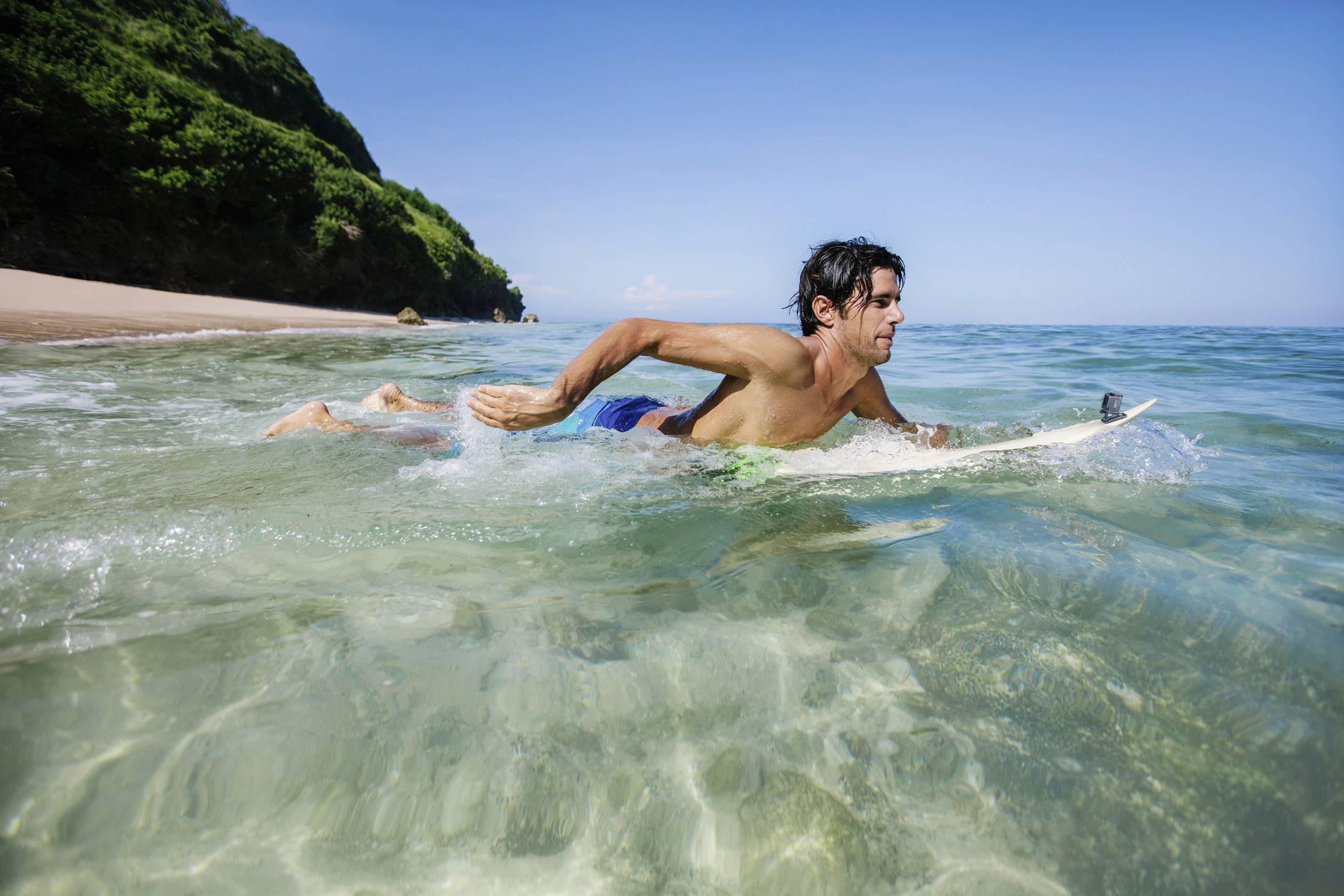Un homme est allongé sur une planche de surf dans l'eau peu profonde, pagayant en direction de la mer. La plage et des collines verdoyantes sont visibles en arrière-plan.