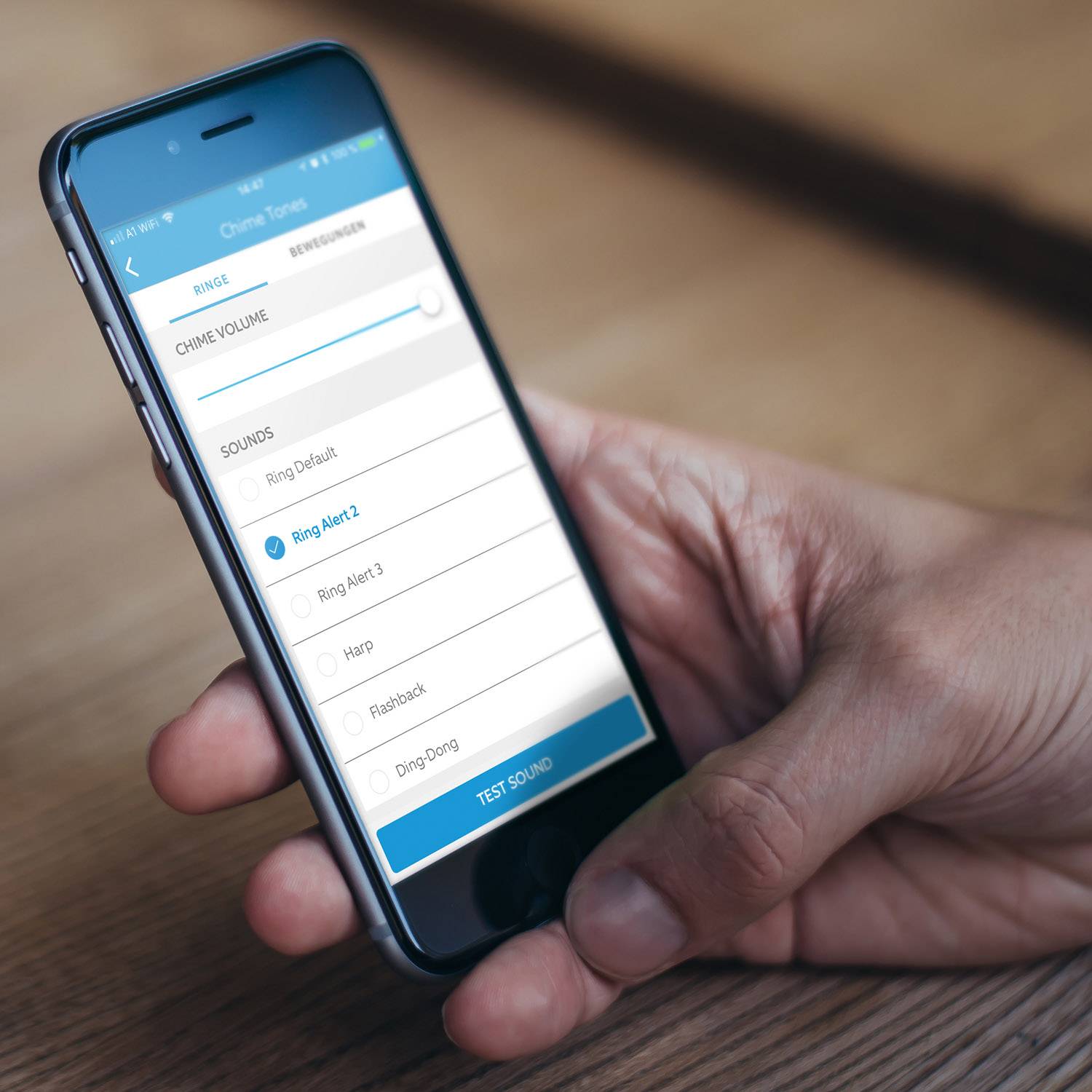 Une personne tient un smartphone avec l'écran ouvert pour sélectionner une sonnerie. Les options comprennent 'Ring Alert 2', 'Harpe' et d'autres.