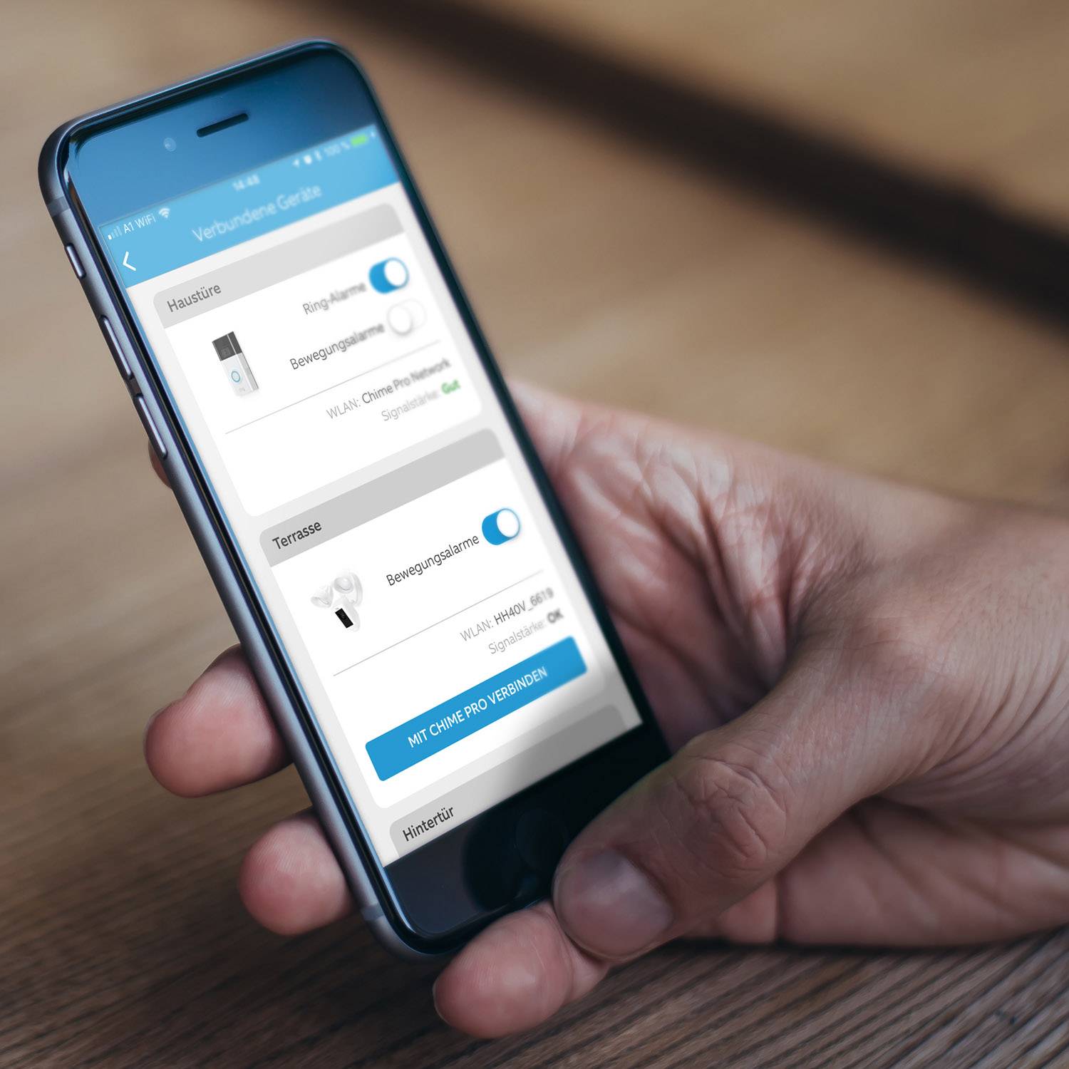 Une personne tient un smartphone avec une application ouverte affichant les paramètres d'appareils domestiques, incluant l'éclairage et le contrôle de la température.