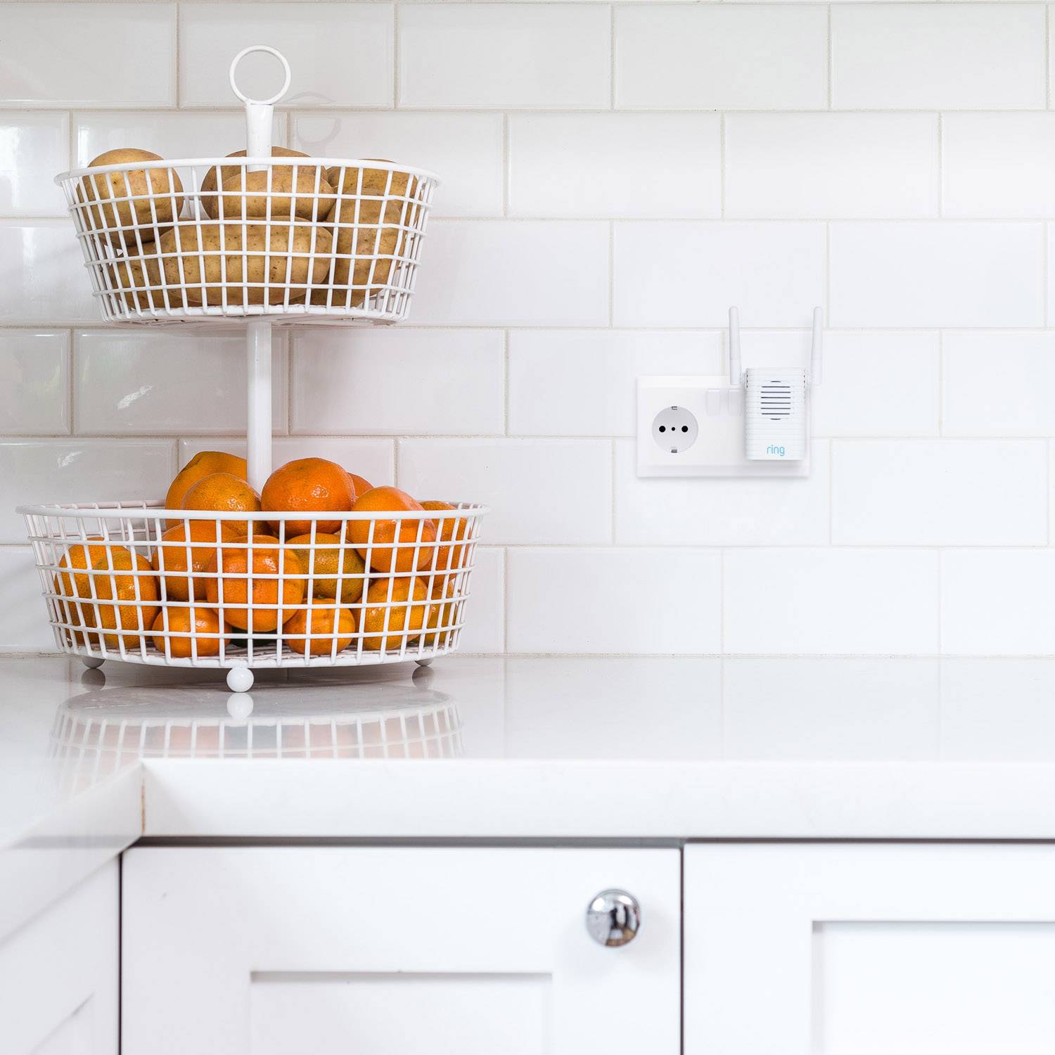 Corbeille à fruits à deux étages posée sur un plan de travail blanc de cuisine, remplie d'oranges et de pommes de terre, devant un mur carrelé de blanc.
