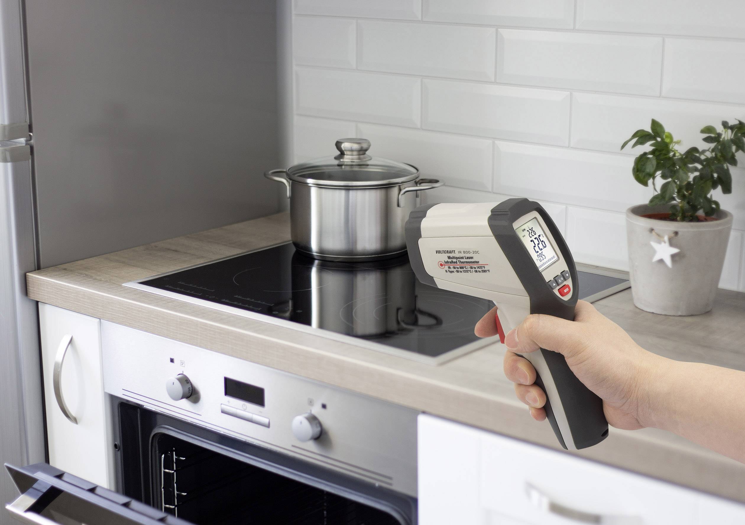 Une personne tient un thermomètre infrarouge dans la cuisine et le pointe vers une casserole sur la cuisinière. L'écran affiche 21,8°C.