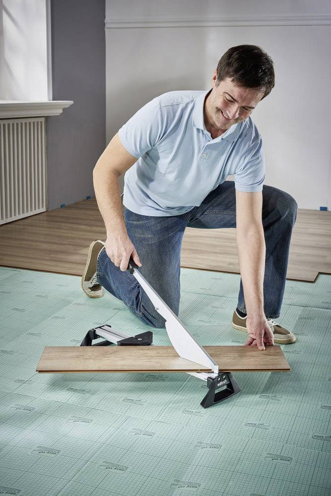 Un homme vêtu de bleu pose un sol stratifié tout en découpant une planche à l'aide d'un outil de coupe.
