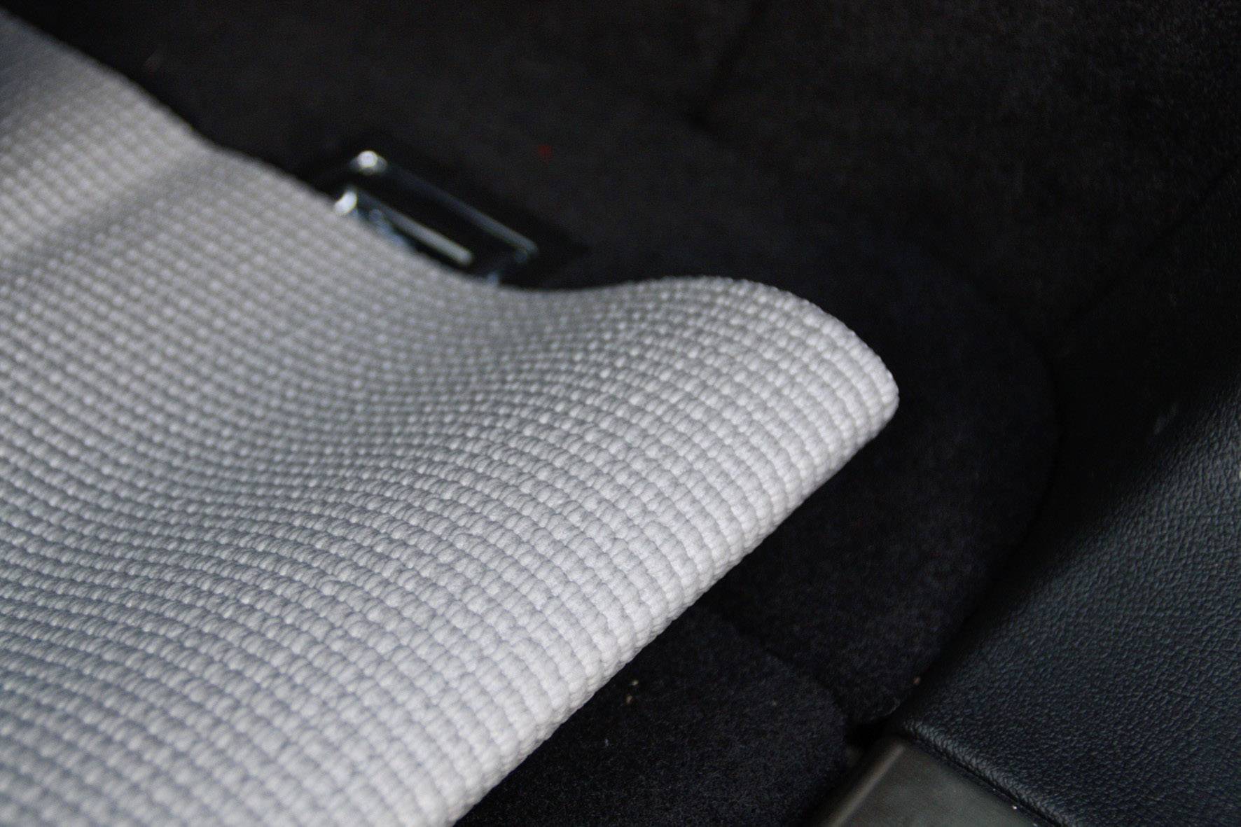 Un tapis gris est posé sur le plancher sombre d'une voiture, partiellement plié. En dessous, une boucle métallique brillante est visible.