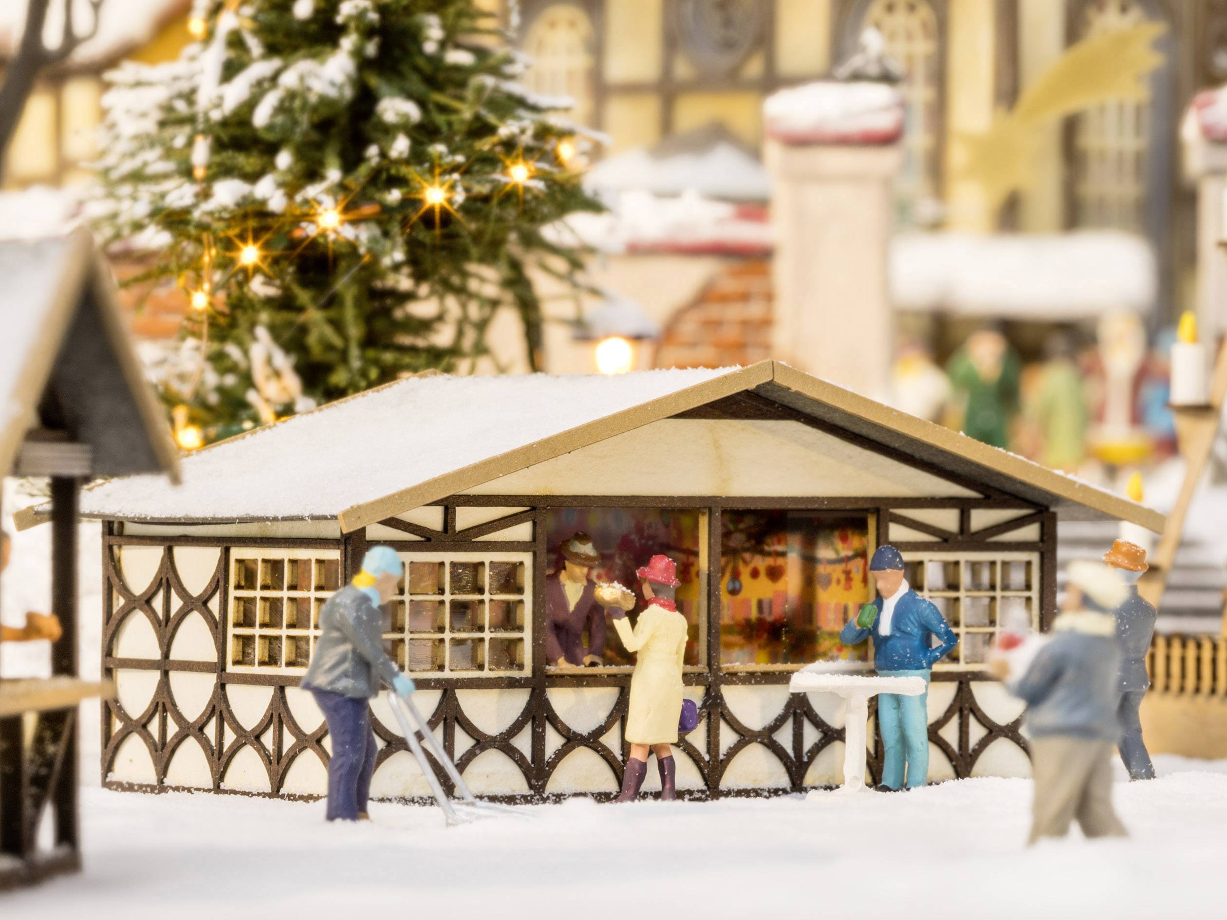 Scène de marché de Noël miniature avec un stand en bois décoré, de la neige et plusieurs personnages. Ils achètent et vendent des marchandises. Des bâtiments et des lumières décorés en arrière-plan.