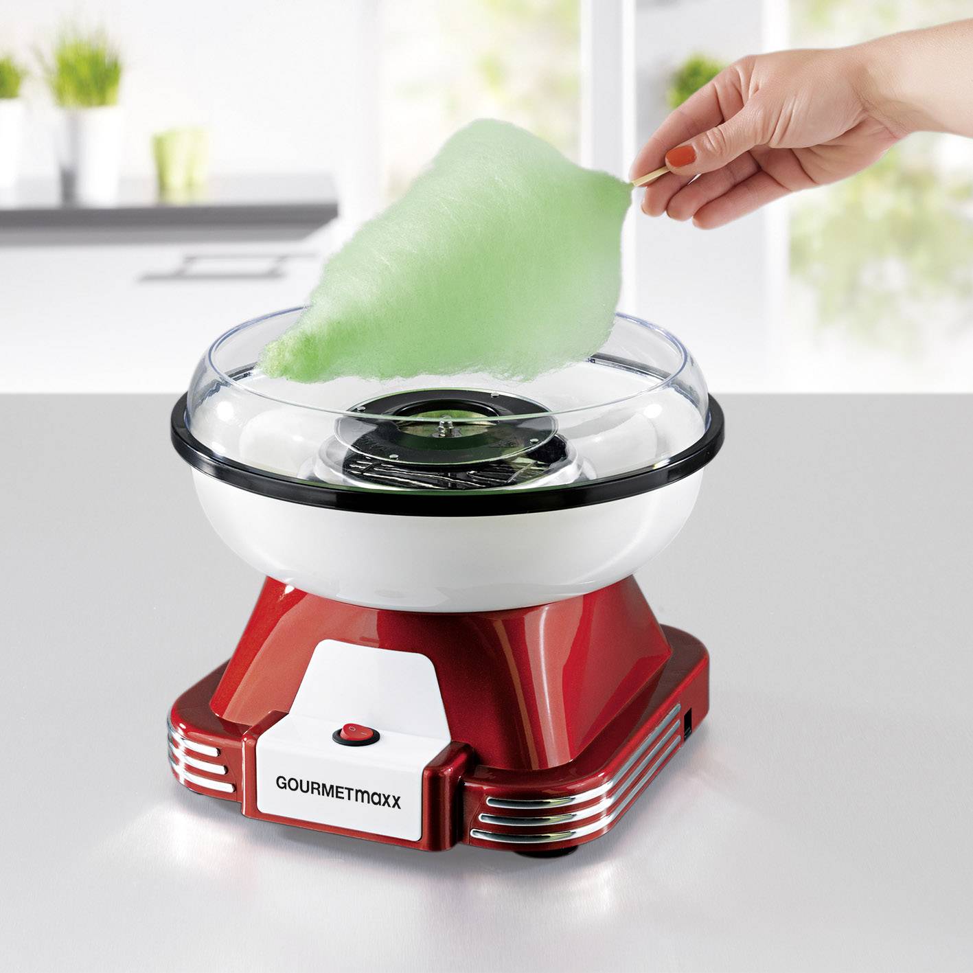 Une main tient de la barbe à papa verte au-dessus d'une machine à barbe à papa rouge dans une cuisine moderne.