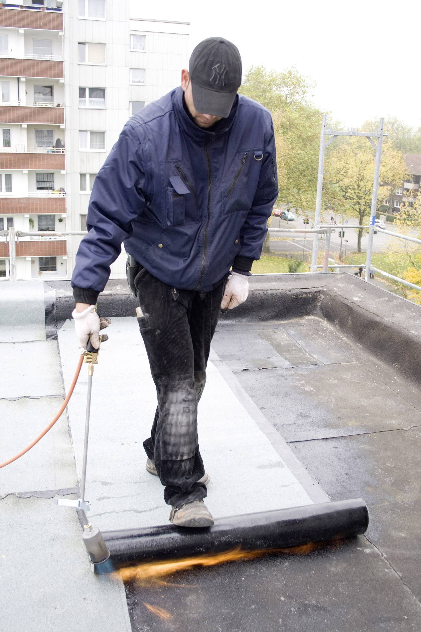Une personne en tenue de travail utilise un chalumeau à gaz pour fixer du matériau de toiture sur un toit plat. Des bâtiments résidentiels sont visibles en arrière-plan.