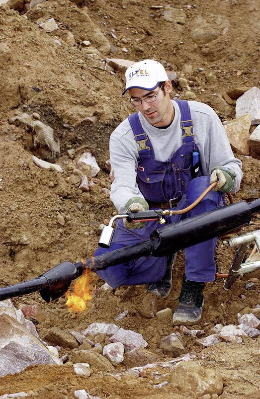 Un ouvrier portant une salopette bleue et une casquette blanche utilise un appareil de soudure pour travailler un tube dans un environnement terreux.