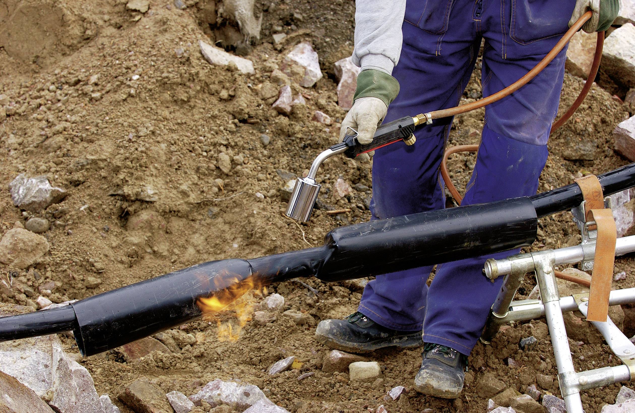 Un travailleur chauffe un grand câble avec un chalumeau à l'extérieur, entouré de terre et de pierre.