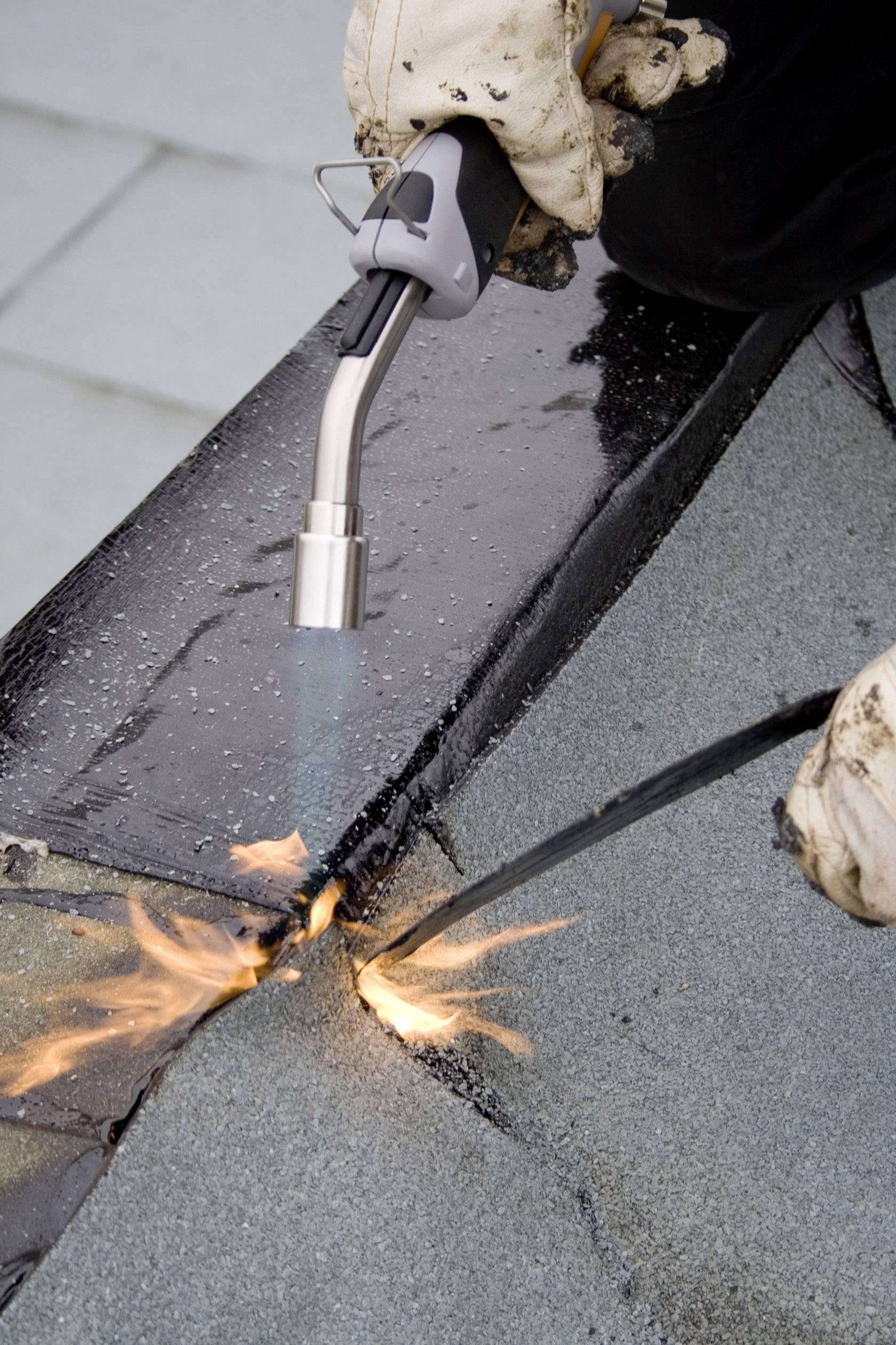 Un ouvrier du bâtiment chauffe des feuilles de bitume avec un chalumeau pour installer une étanchéité de toiture. Des flammes sont visibles lors de la fusion de la surface.