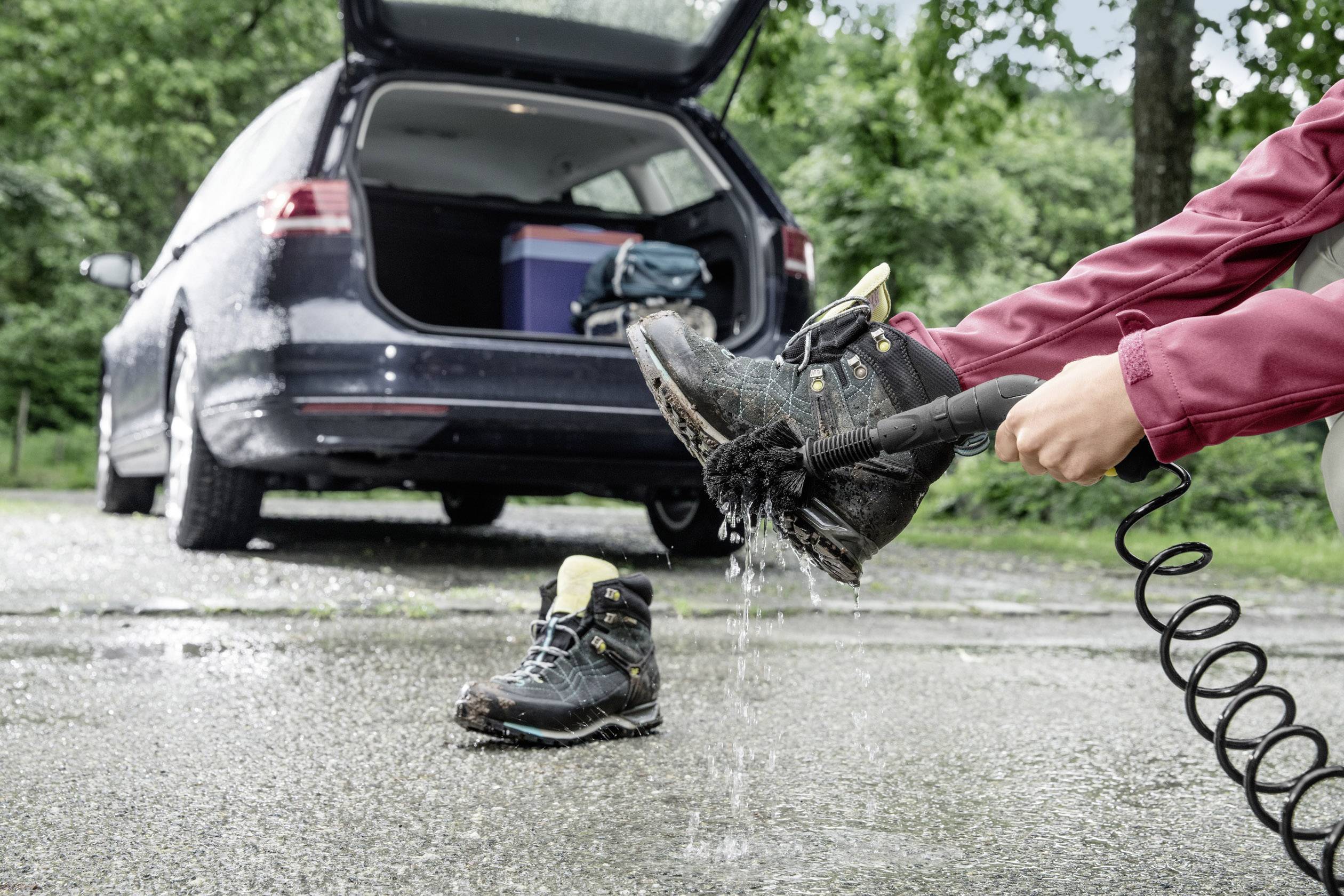 Une personne pulvérise de l'eau sur des chaussures de randonnée à côté d'une voiture garée, dont le coffre est ouvert et rempli d'équipement. Activité de plein air ou préparation de voyage.