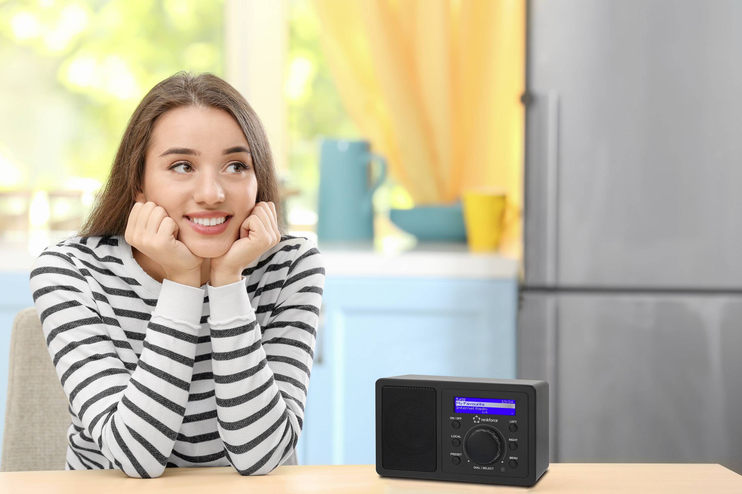 Une femme souriante portant un pull rayé est assise à table à côté d'une radio numérique dans une cuisine lumineuse.